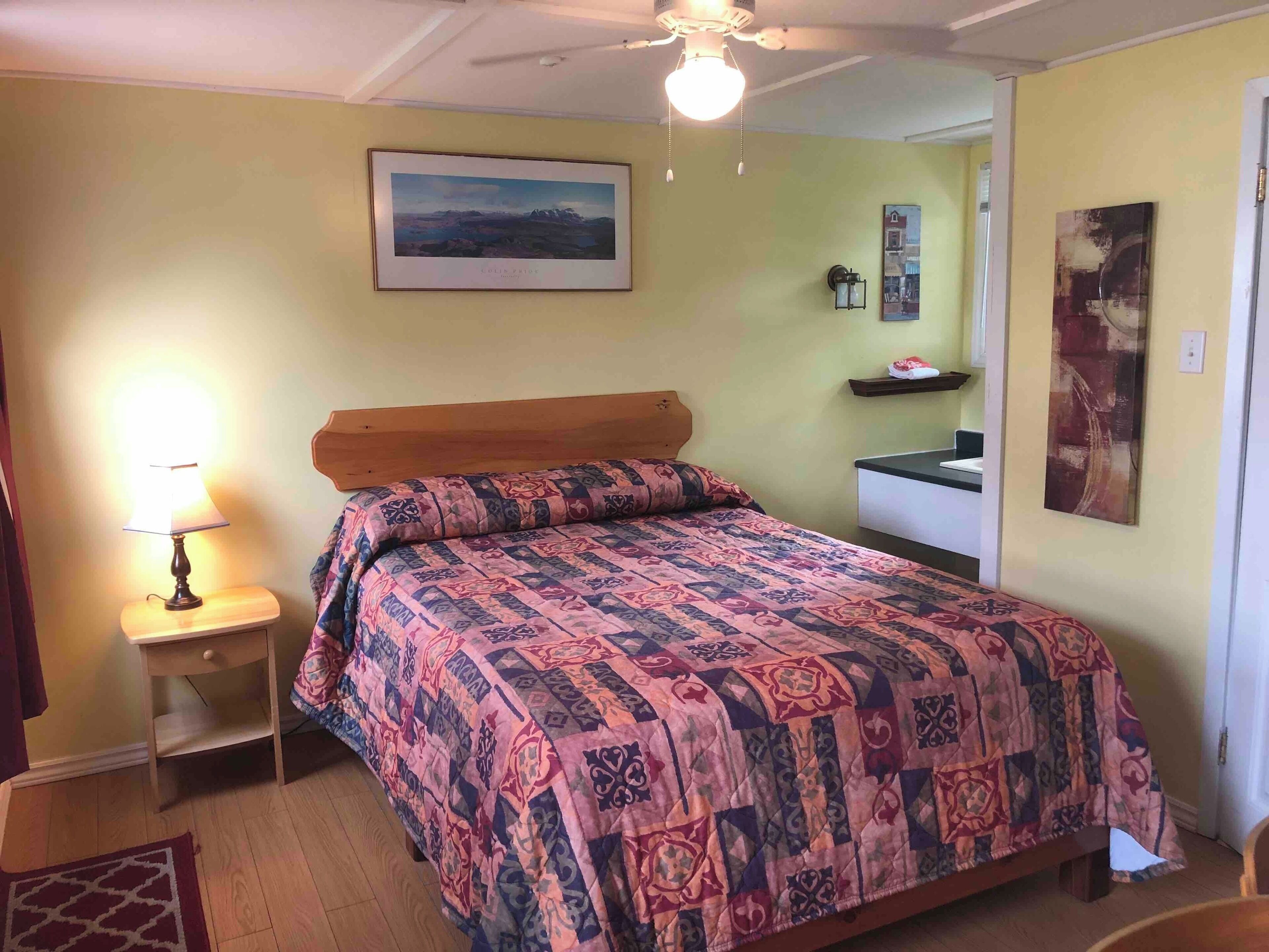 Una camera da letto con un letto, un comodino, una lampada, una ventola a soffitto, un quadro appeso al muro e una piccola mensola con oggetti.