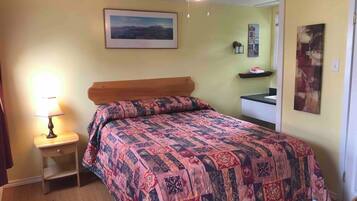 Una camera da letto con un letto, un comodino, una lampada, una ventola a soffitto, un quadro appeso al muro e una piccola mensola con oggetti.