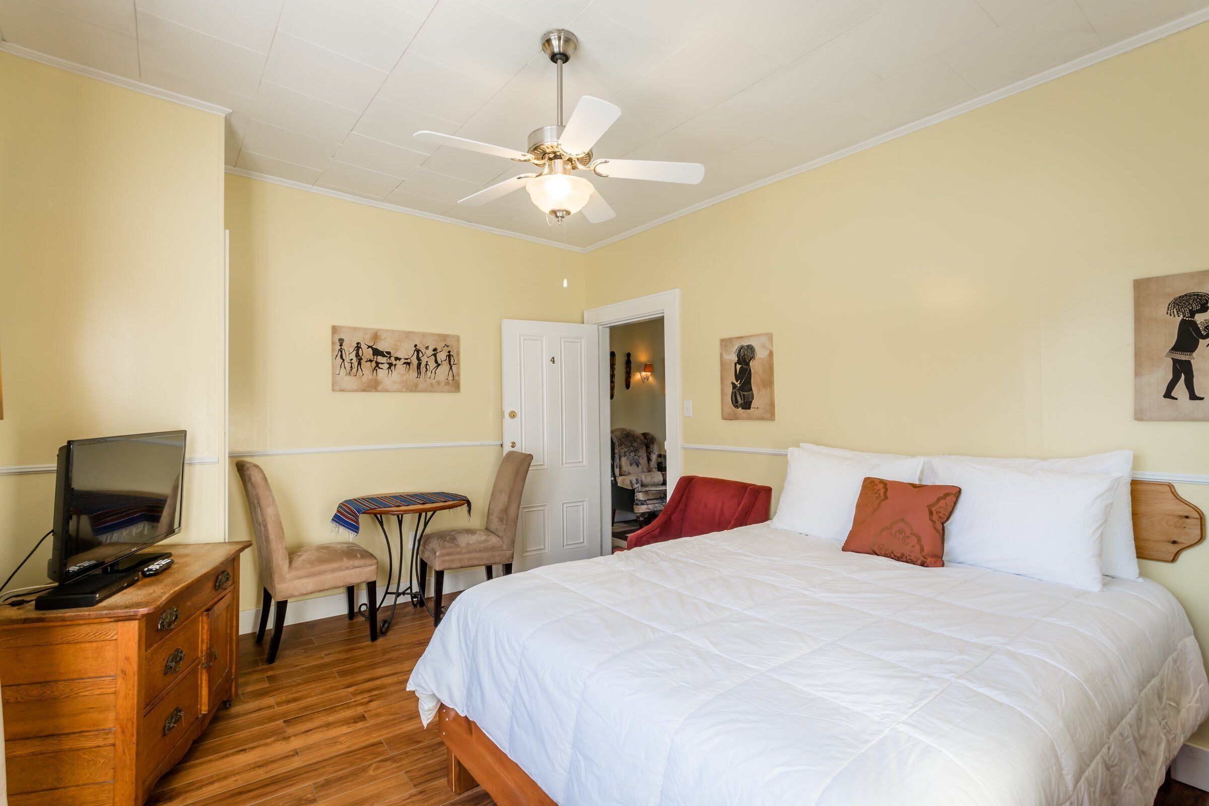 Une chambre à coucher avec un lit, un téléviseur posé sur une commode en bois, une chaise et un ventilateur de plafond.