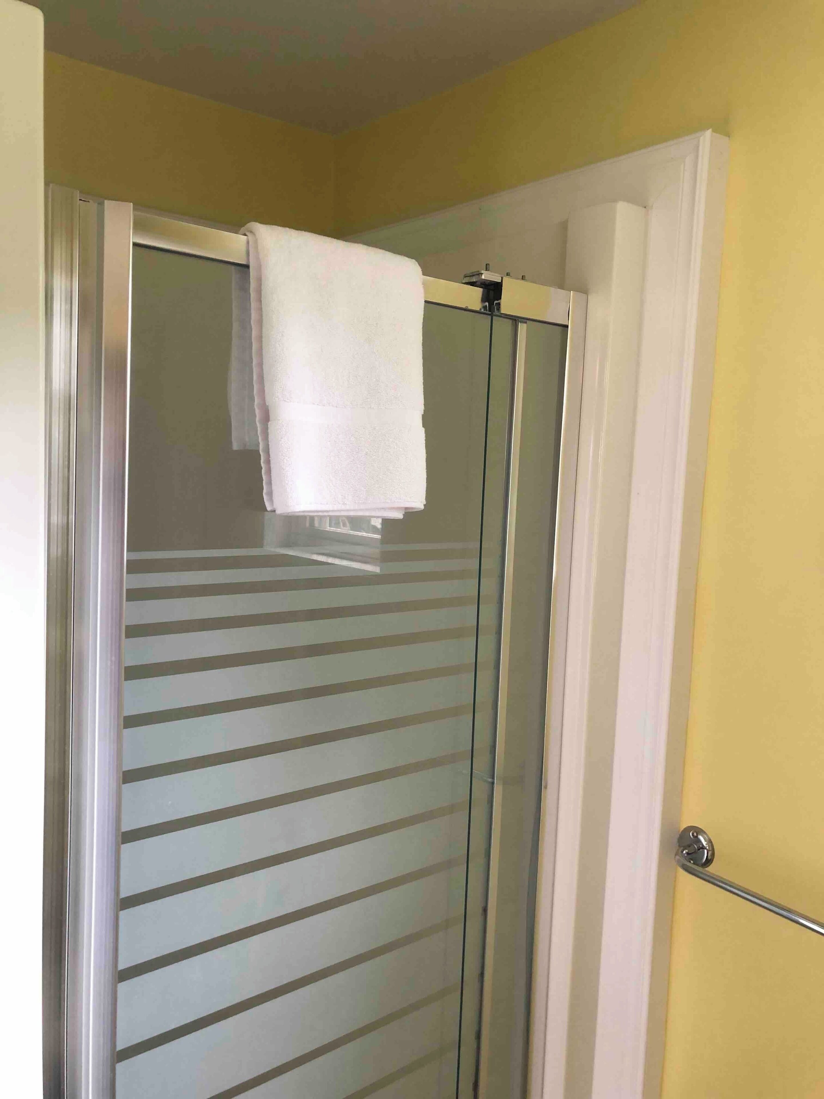 A bathroom with a glass shower door, a white towel hanging on the door handle, and a yellow wall.