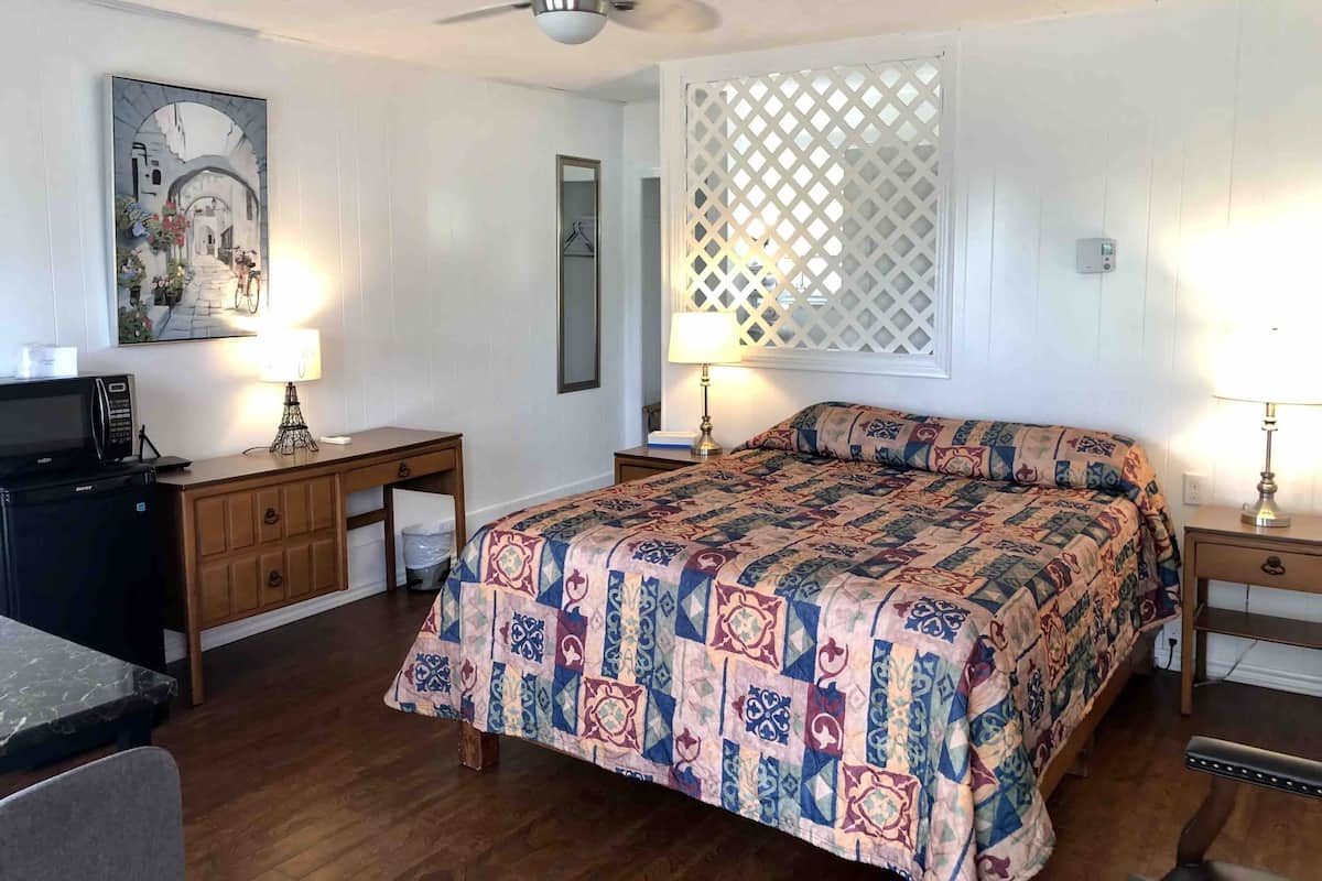 Une chambre à coucher avec un lit, deux tables de chevet avec des lampes, un bureau avec un ordinateur, un ventilateur de plafond et un tableau accroché au mur.