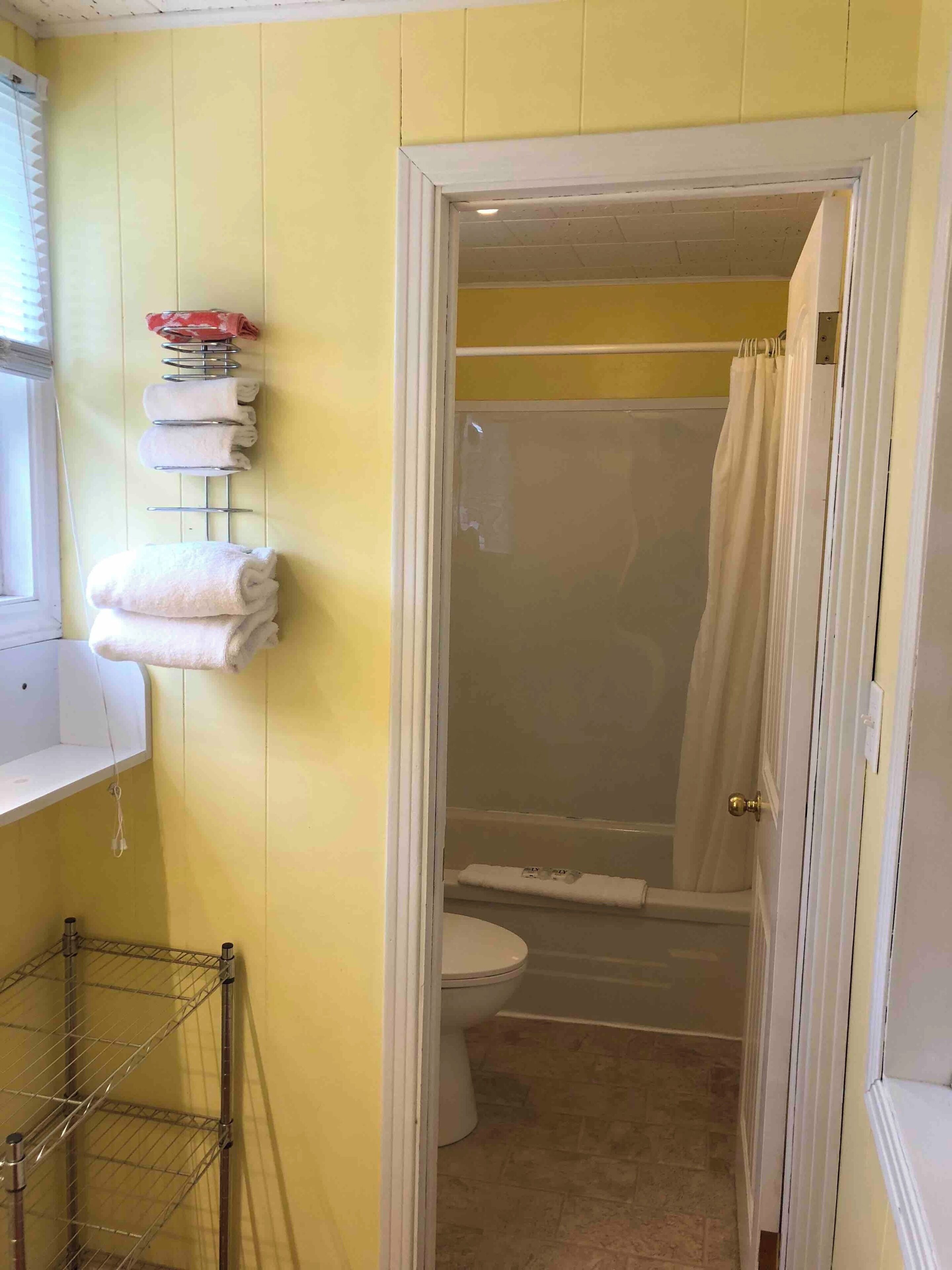 A bathroom with yellow walls, a white door, a shower curtain, a toilet, and a metal drying rack with towels.