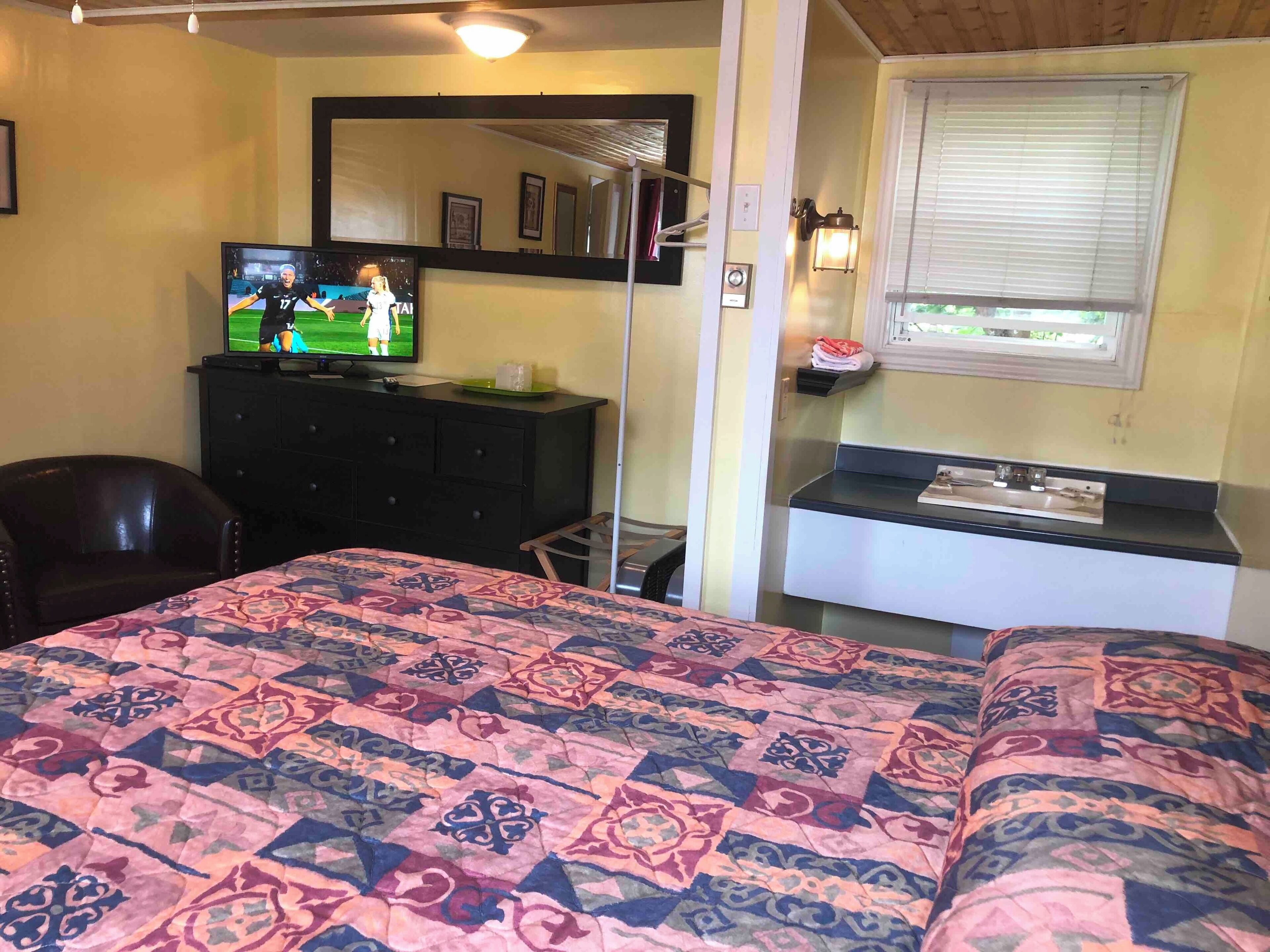 Ein Schlafzimmer mit einem Bett, einem Fernseher auf einer Kommode, einem Sessel und einem Fenster mit Jalousien.
