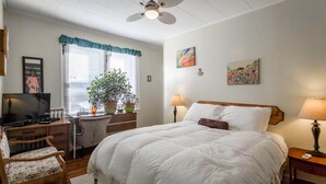 Een slaapkamer met een bed, een bureau, een stoel, een televisie, een plafondventilator en twee schilderijen aan de muur.