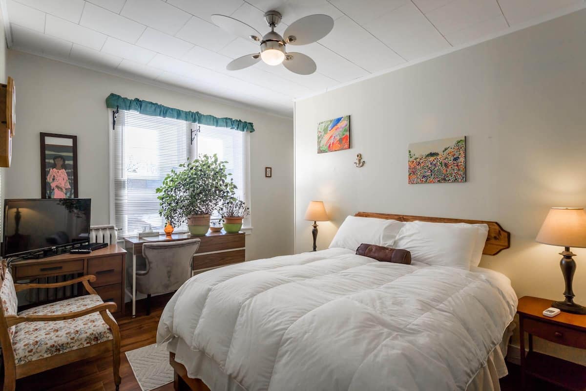 Une chambre à coucher comprenant un lit, un bureau, une chaise, un téléviseur, un ventilateur de plafond et deux tableaux accrochés au mur.