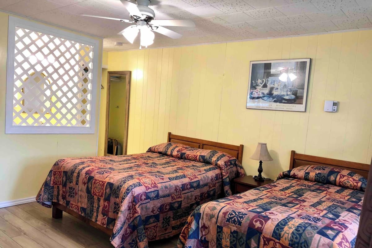 Une chambre avec deux lits, un ventilateur de plafond, une fenêtre ornée d’un motif en treillis et un tableau encadré au mur.