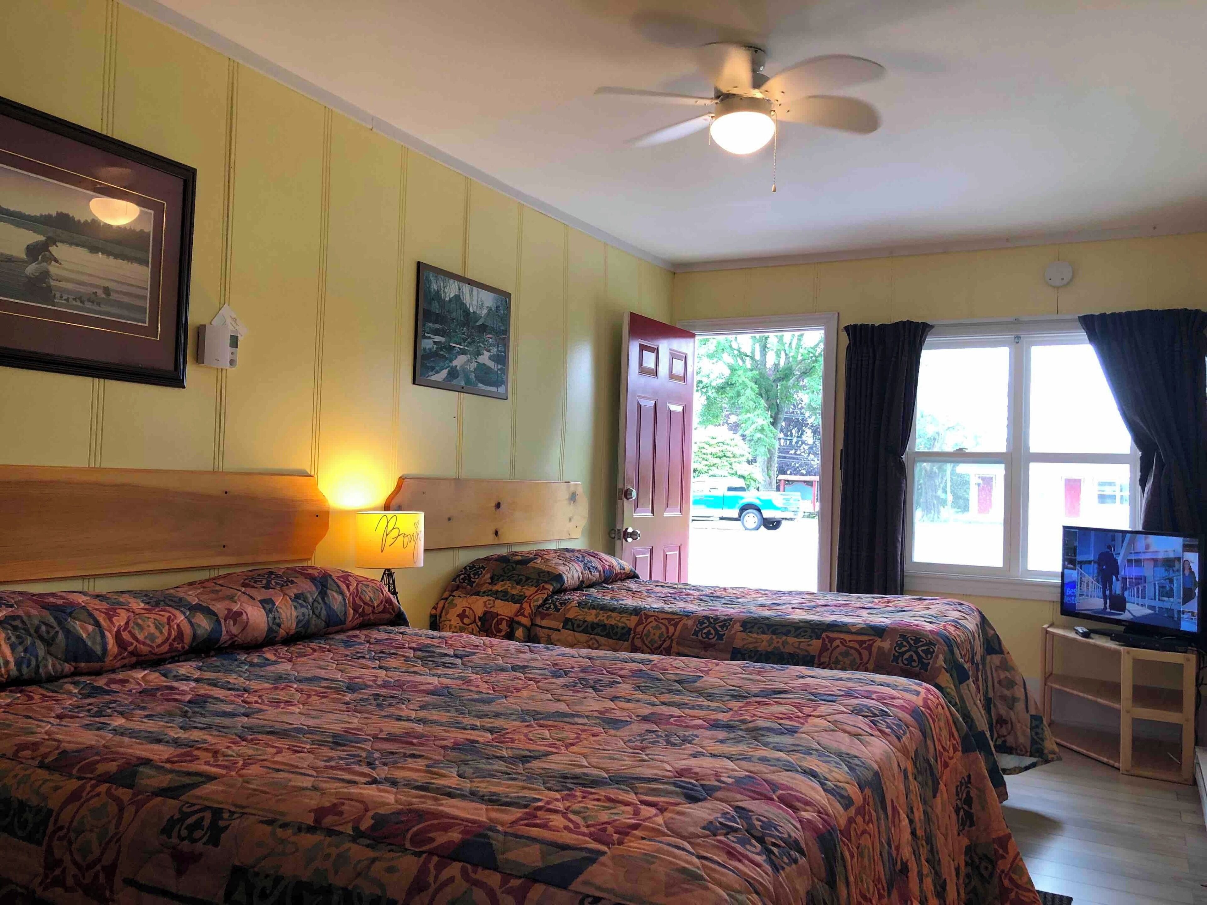 A hotel room with two beds, a ceiling fan, a TV on a stand, and a window with curtains.