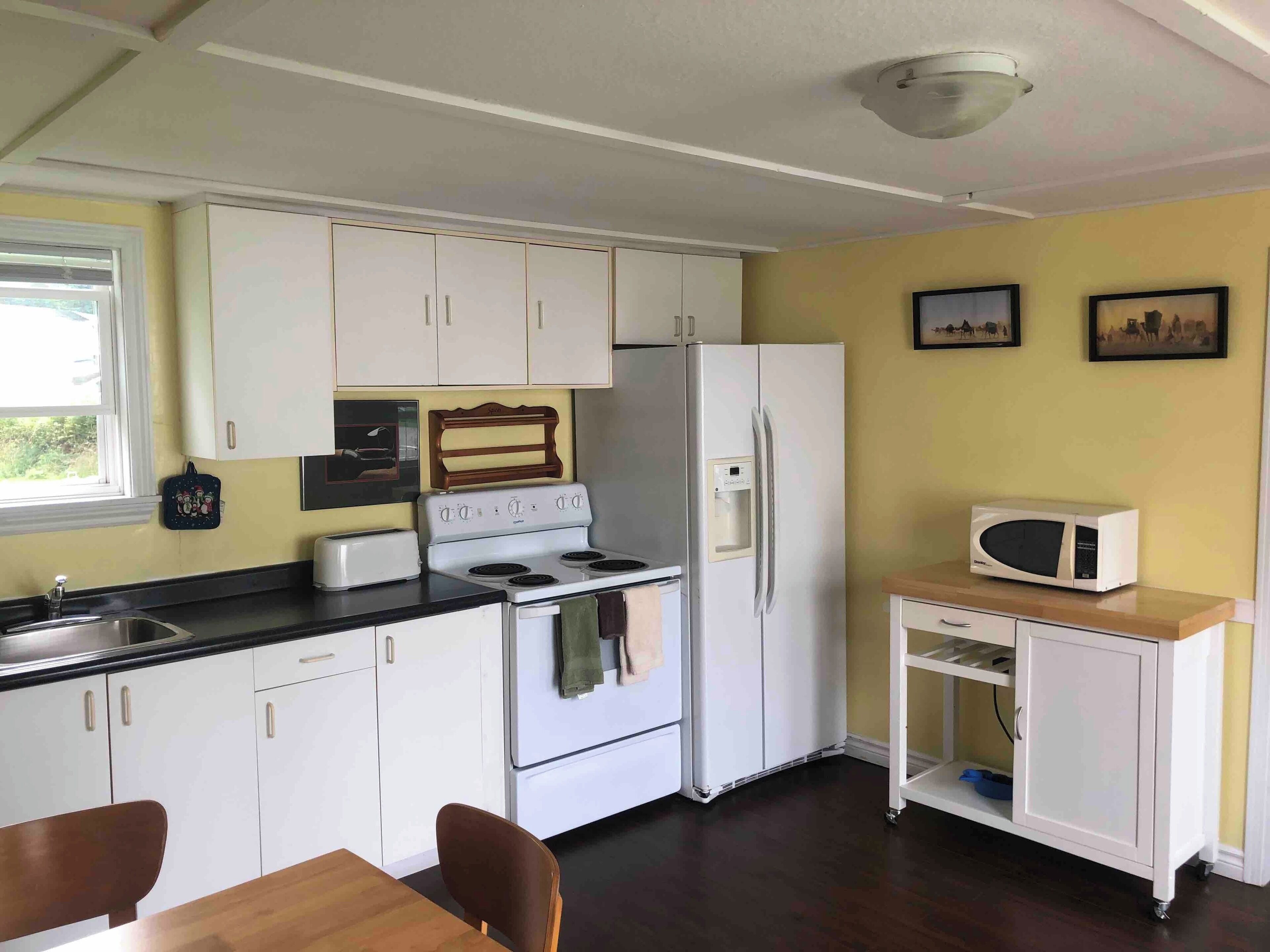 A compact kitchen with white cabinets, a stove, a refrigerator, and a microwave. There is a window providing natural light and a small shelf unit with a microwave on top.