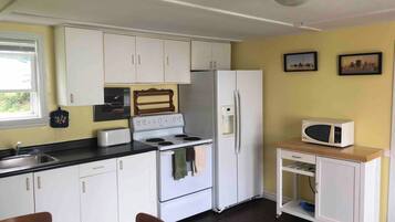 A compact kitchen with white cabinets, a stove, a refrigerator, and a microwave. There is a window providing natural light and a small shelf unit with a microwave on top.