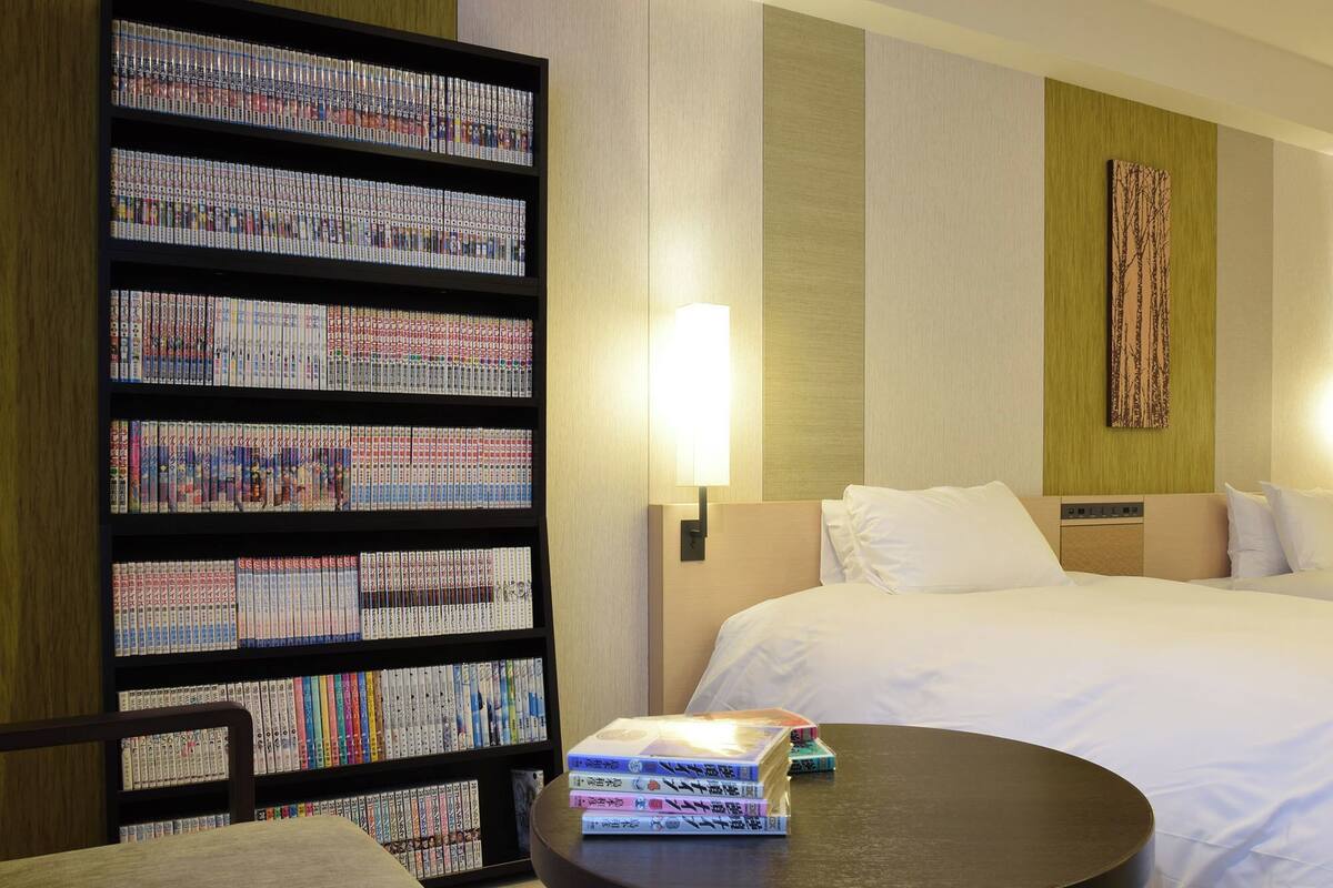 A hotel room with a large bookshelf filled with books, a neatly made bed, a round table with a few books, and a wall-mounted lamp.