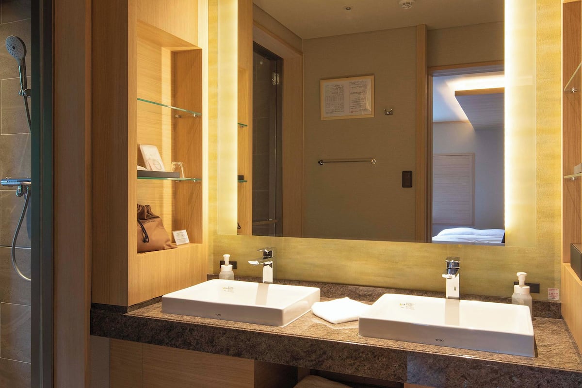 A modern bathroom with a glass shower enclosure, wooden vanity, and a large mirror reflecting a door and a bed.