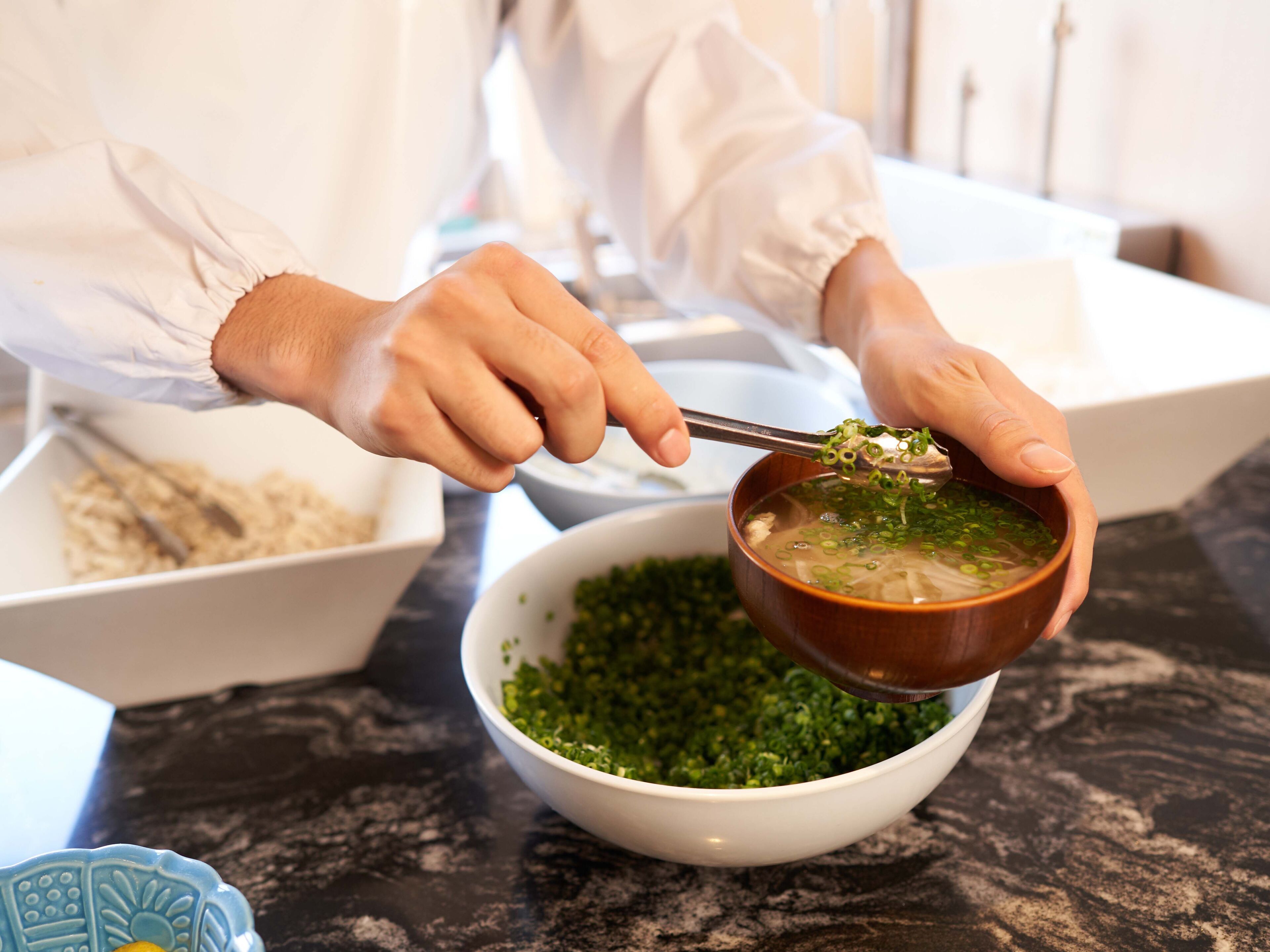 A person preparing a bowl of soup with chopsticks, with ingredients and bowls on a kitchen counter.