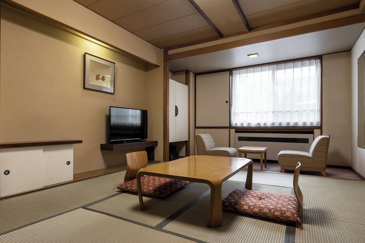 A traditional Japanese-style room with tatami flooring, a low table, two chairs, a television, and a large window with curtains.