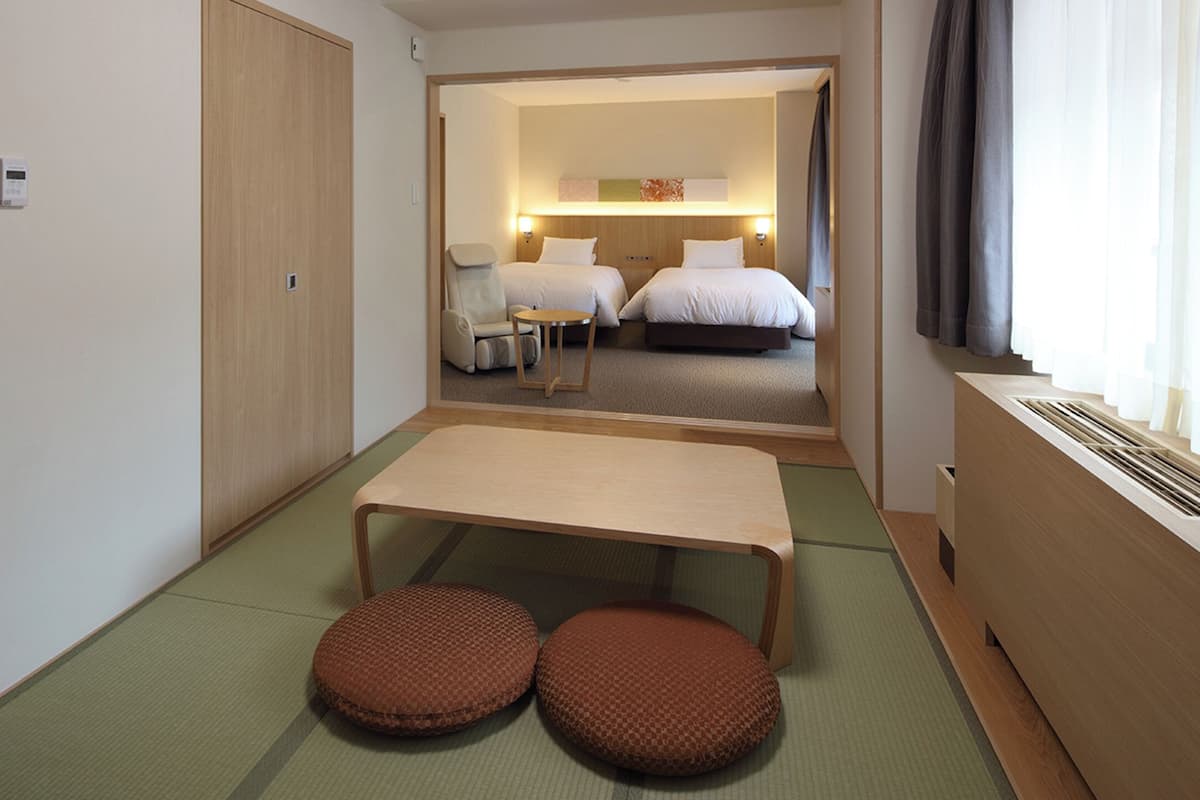 A hotel room with a bed, a small table, two ottomans, a radiator, and a window with curtains.
