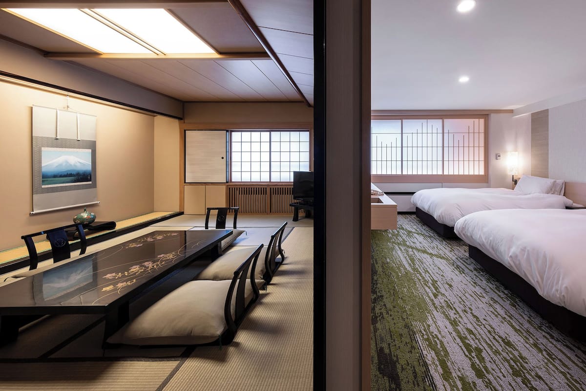 A hotel room with a traditional tatami mat seating area, a bed with white linens, and a view of the cityscape through large windows.
