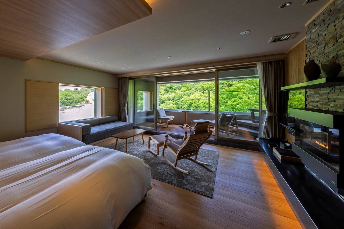 A modern hotel room with a large bed, a seating area with a table and chairs, a fireplace, and a balcony with a view of greenery.