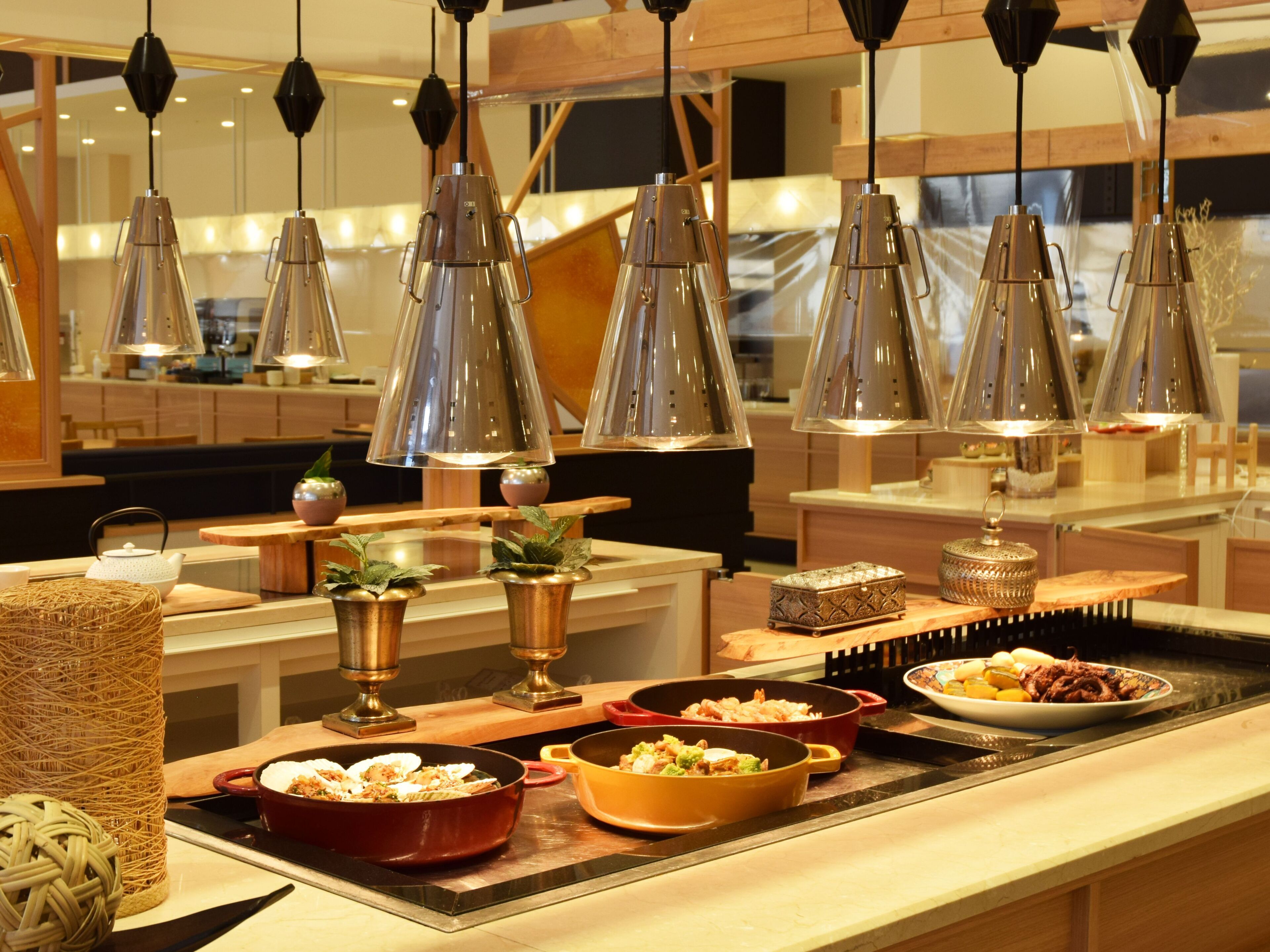 A buffet setup with various dishes, including a red pan and a yellow pan, and decorative items like a woven basket and a small plant.