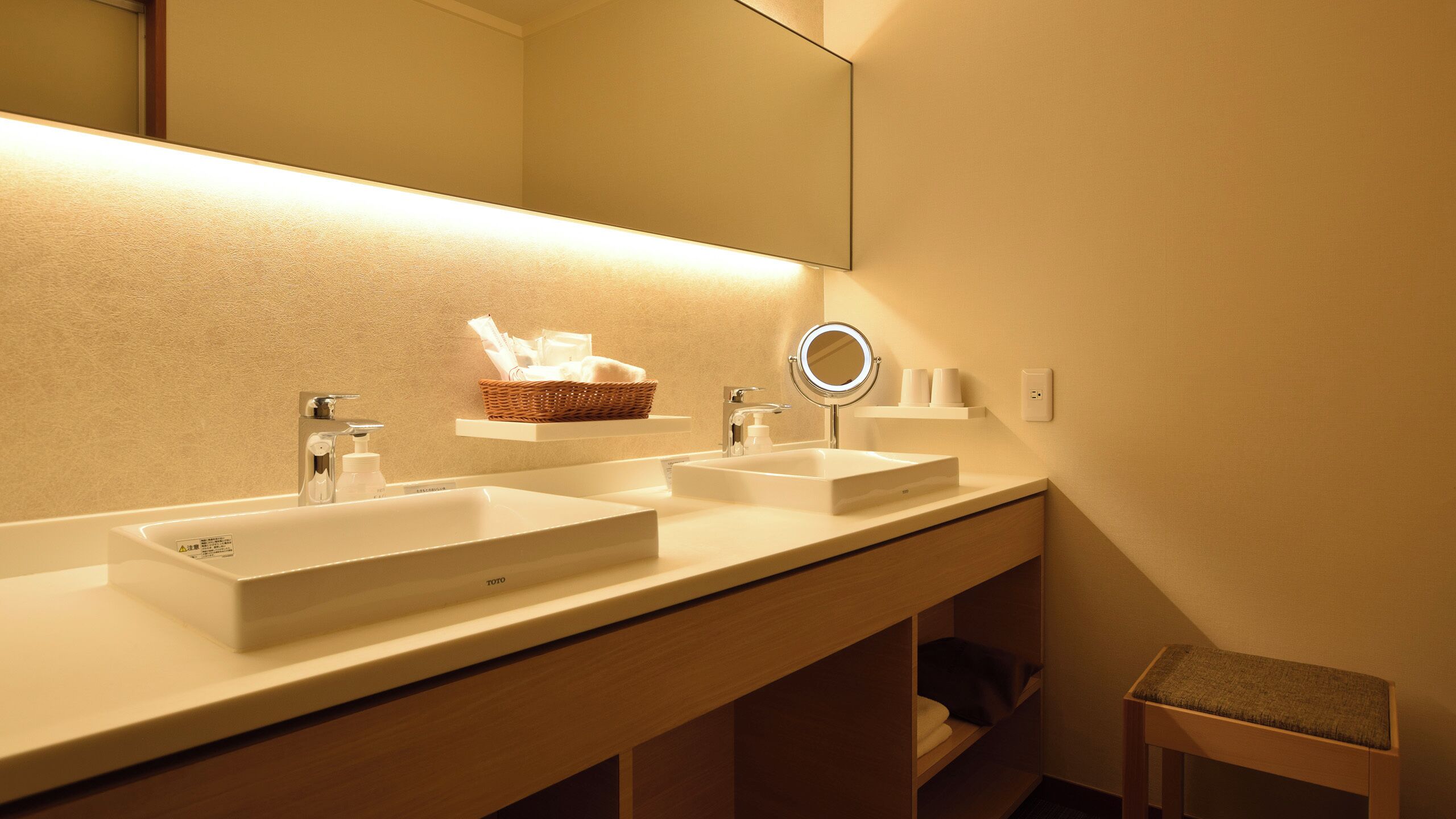A hotel bathroom with two sinks, a mirror, a basket with toiletries, and a small stool.