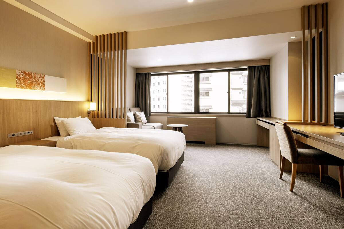 A hotel room with two beds, a desk, and a window with a view of buildings.