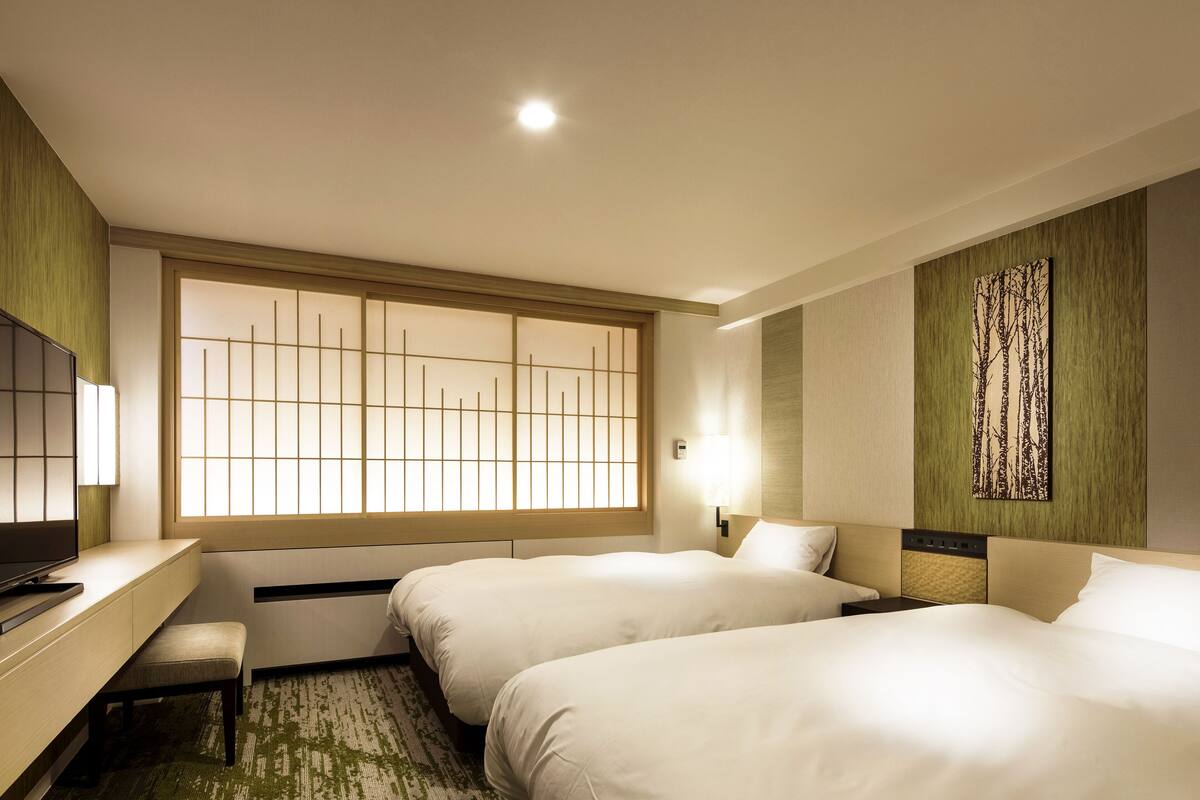 A hotel room with two beds, a desk, a bench, a TV, a large window with a view, and a wall-mounted artwork.