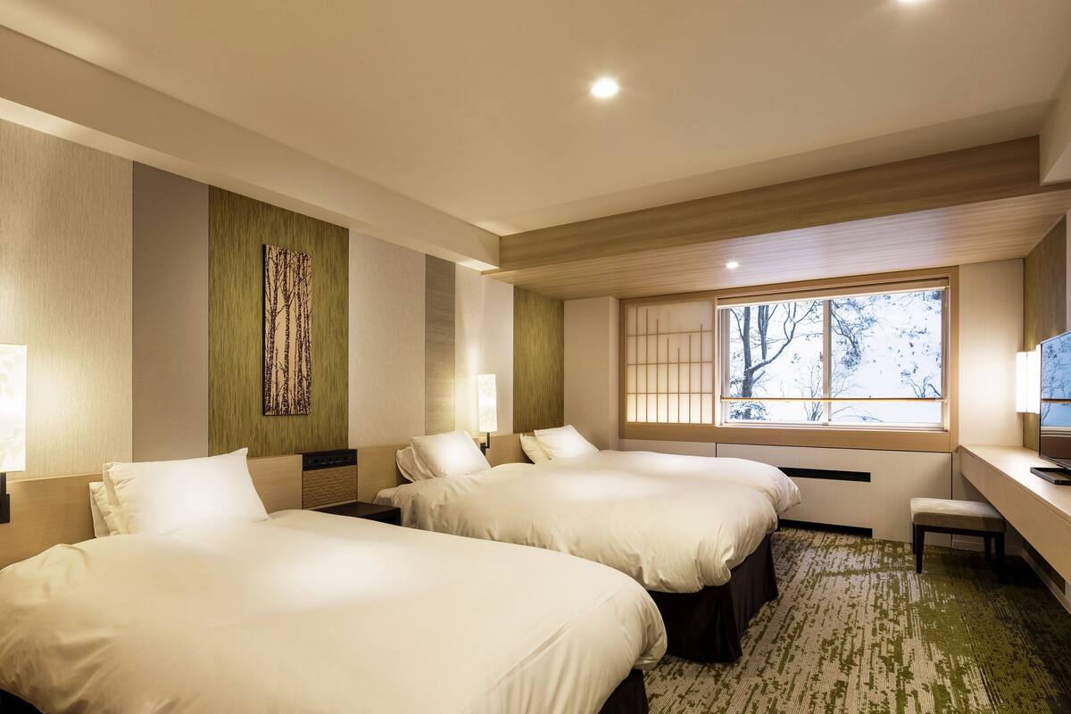 A hotel room with two beds, a desk, a chair, a window with a view of trees, and a wall-mounted artwork.