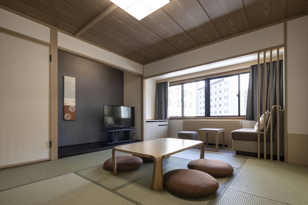 A room with tatami flooring, a low table, cushions, a television, and a window with curtains.