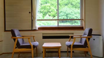 Una habitación con dos sillas de madera y una mesita, una ventana con vistas a árboles verdes, y un radiador en la pared.