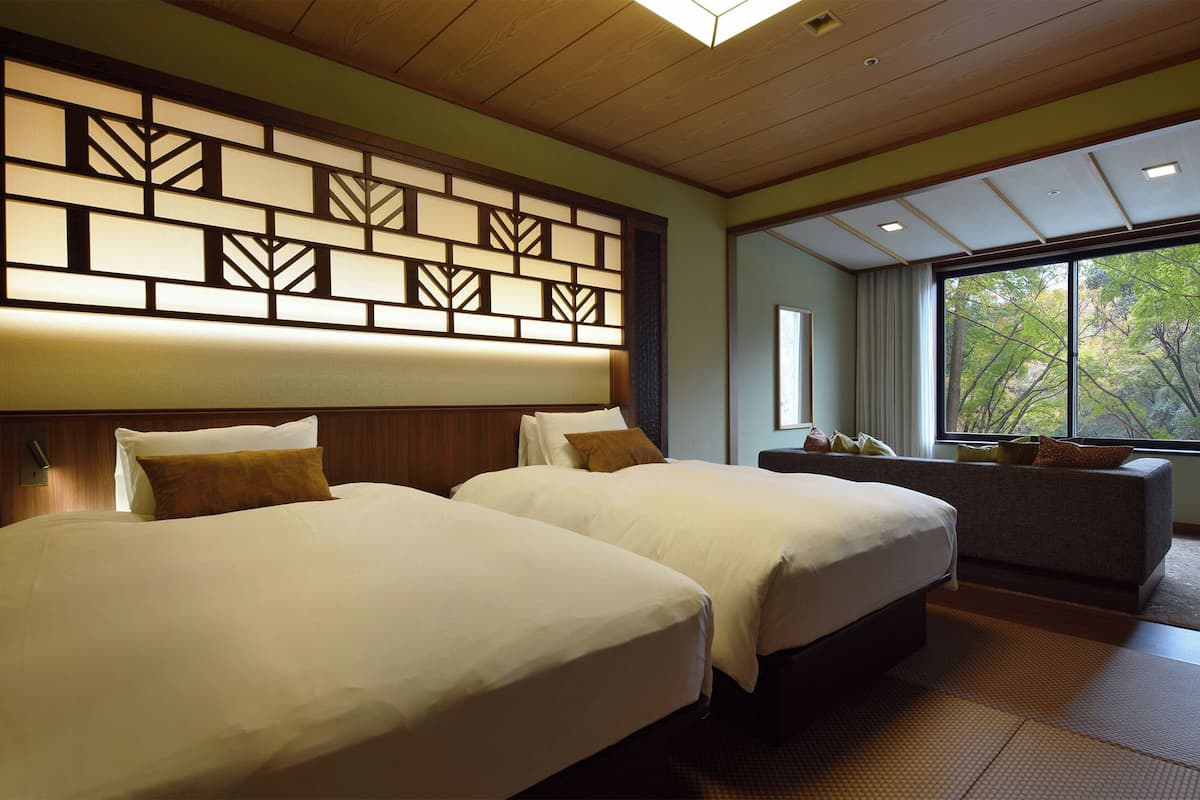 A hotel room with two beds, a sofa, a large window with a view of trees, and a decorative wall panel.