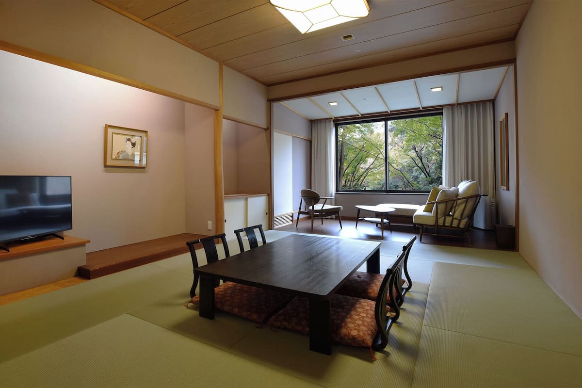 A traditional Japanese-style room with tatami flooring, a low table with cushions, a television, and a large window with a view of trees.