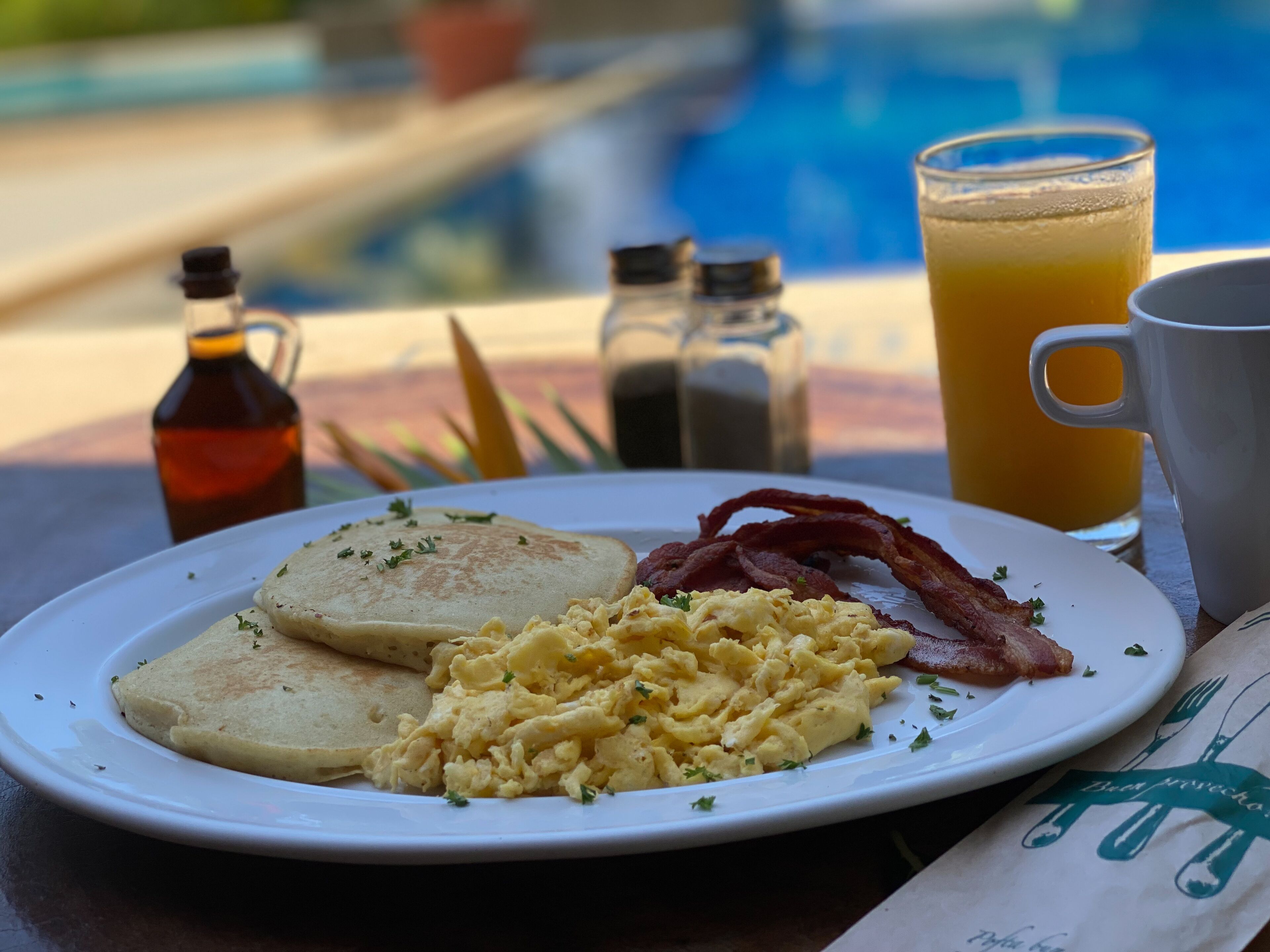A breakfast plate with eggs, bacon, and toast, a glass of orange juice, and a coffee cup on a table by a pool.