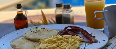 A breakfast plate with eggs, bacon, and toast, a glass of orange juice, and a coffee cup on a table by a pool.