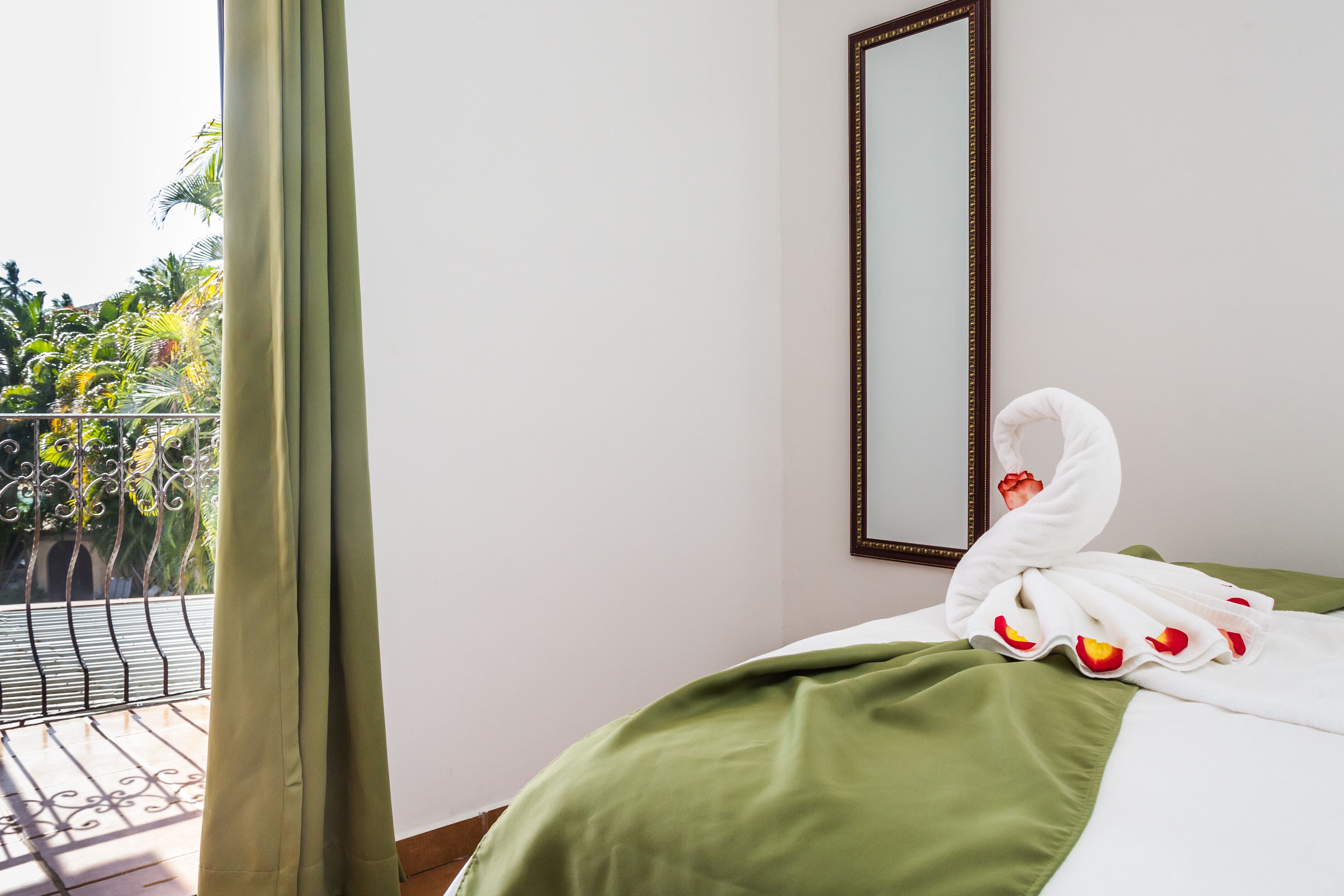 A hotel room with a balcony, a bed with a green bedspread, and a white towel with rose petals on it.