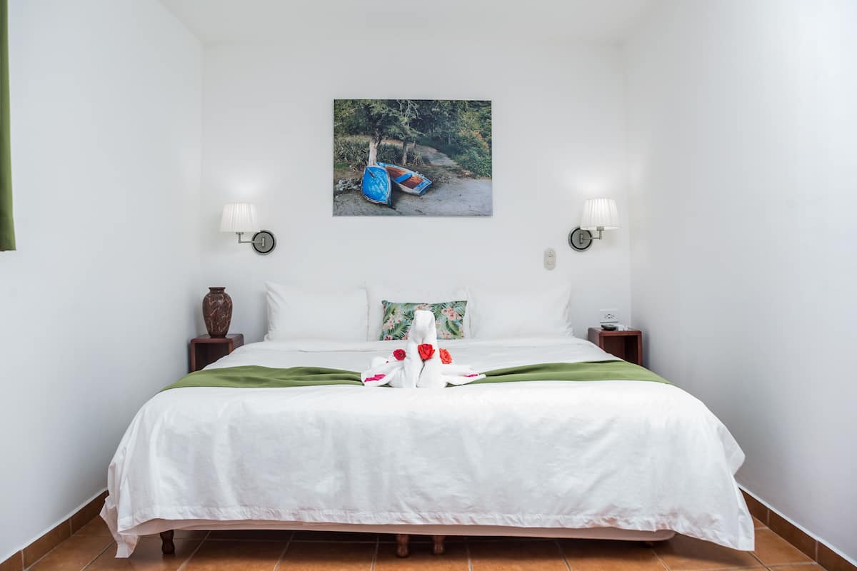A neatly made bed with a white bedspread, two nightstands with lamps, a painting of boats on the wall, and a green throw blanket.