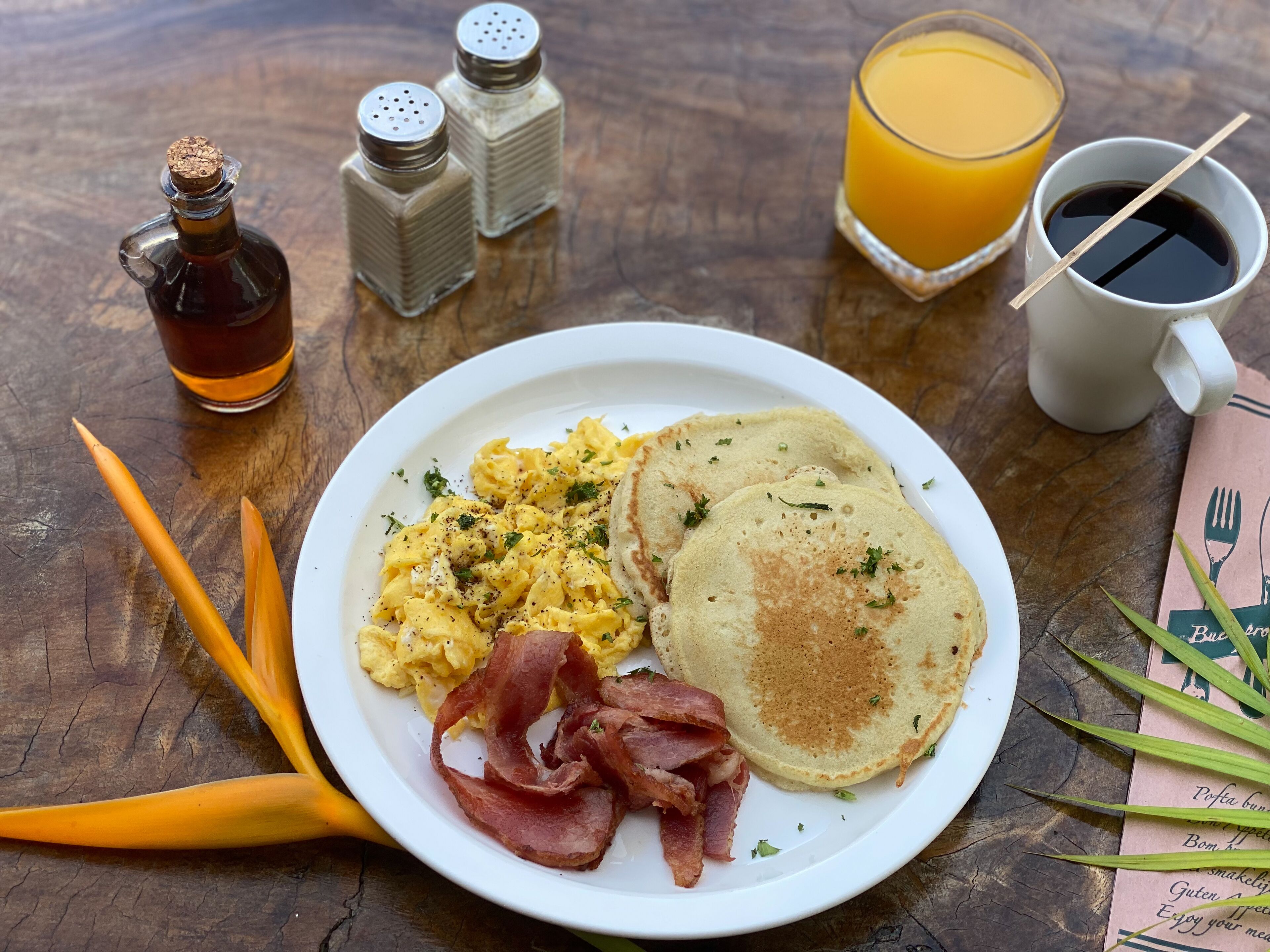 A breakfast plate with scrambled eggs, bacon, and pancakes, accompanied by a glass of orange juice and a cup of coffee, on a wooden table.
