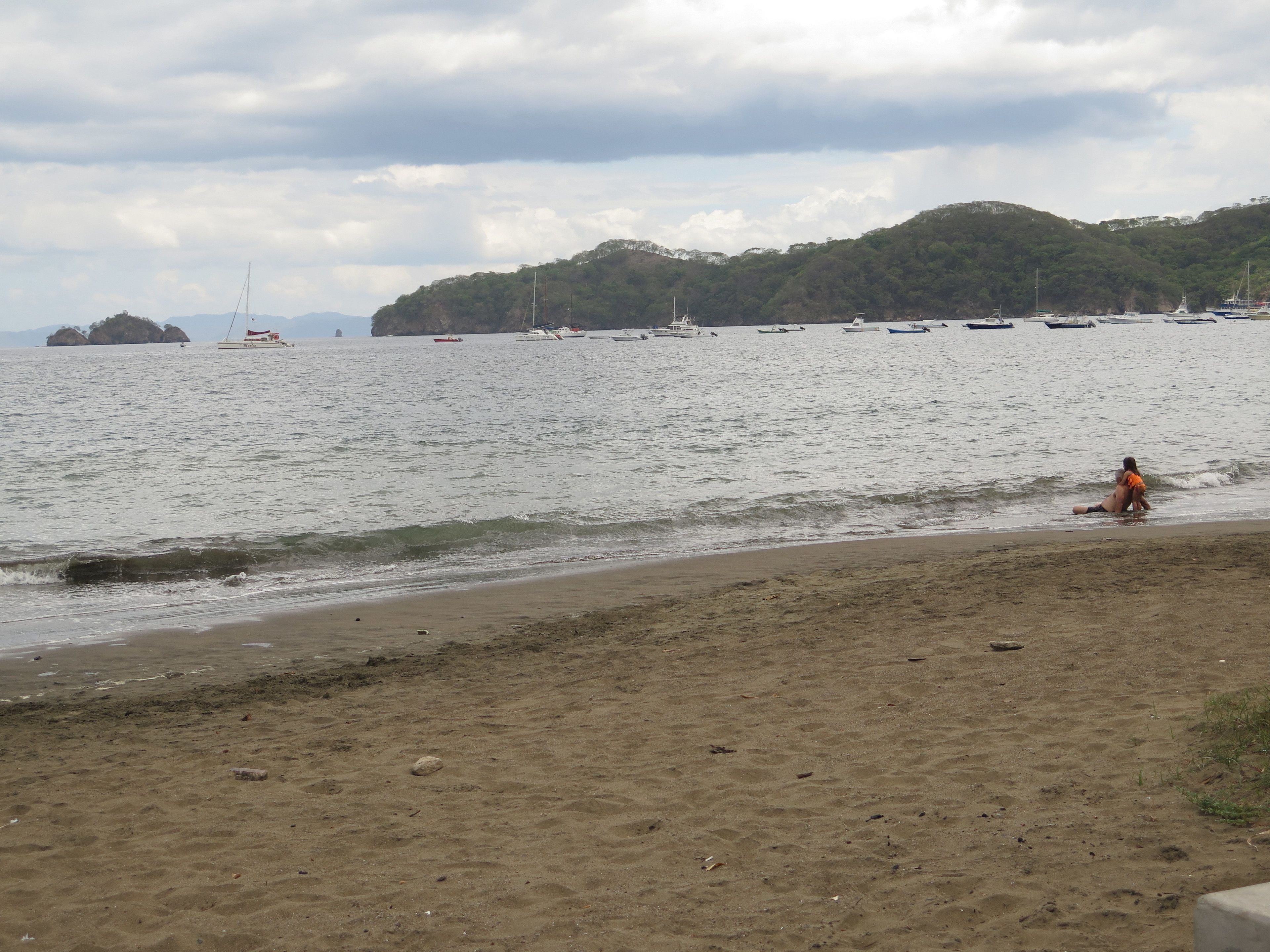 A beach with boats in the water and a person sitting on the sand.