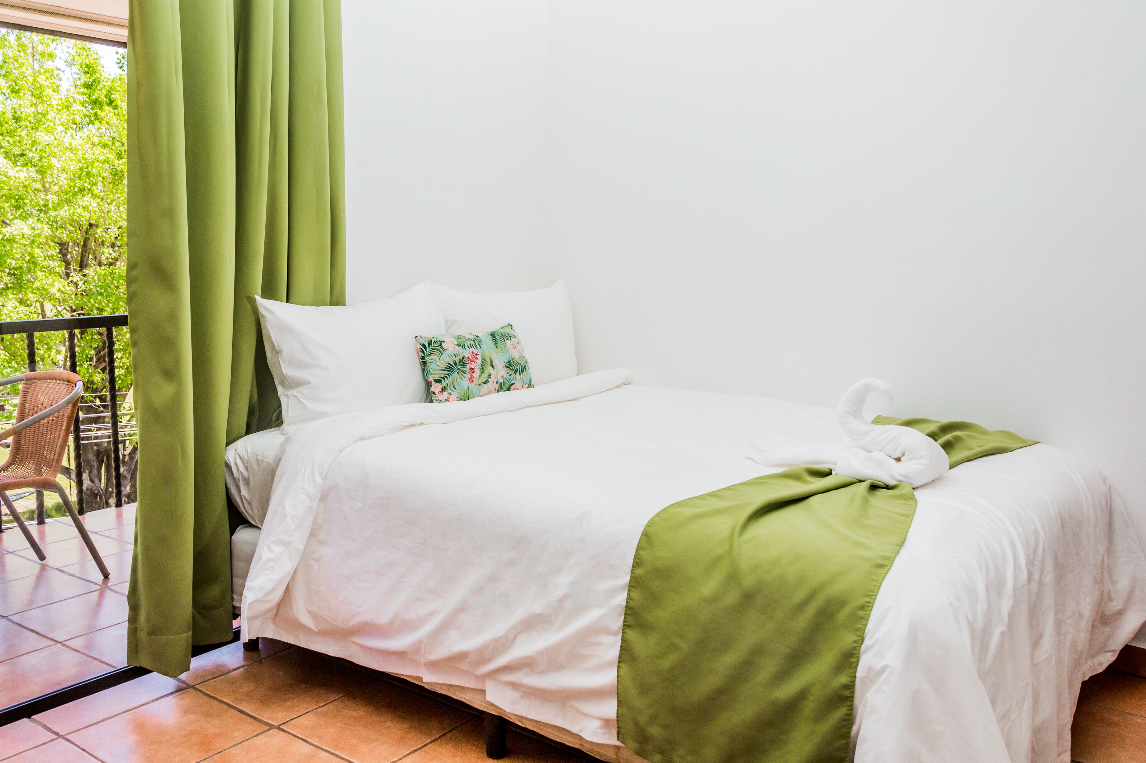 A neatly made bed with white linens and a green accent, a wicker chair, and a view of greenery through a window.