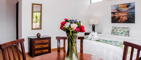 A hotel room with a wooden dining table, a vase of flowers, a bed with a white bedspread, a nightstand, a lamp, and a painting on the wall.