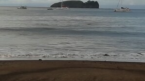 A beach with calm waters, a large rock formation, and sailboats.
