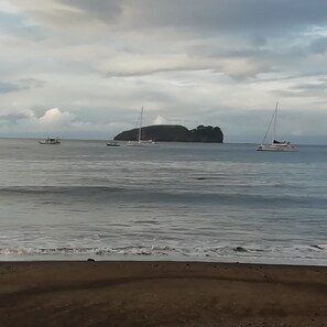 A beach with calm waters, a large rock formation, and sailboats.