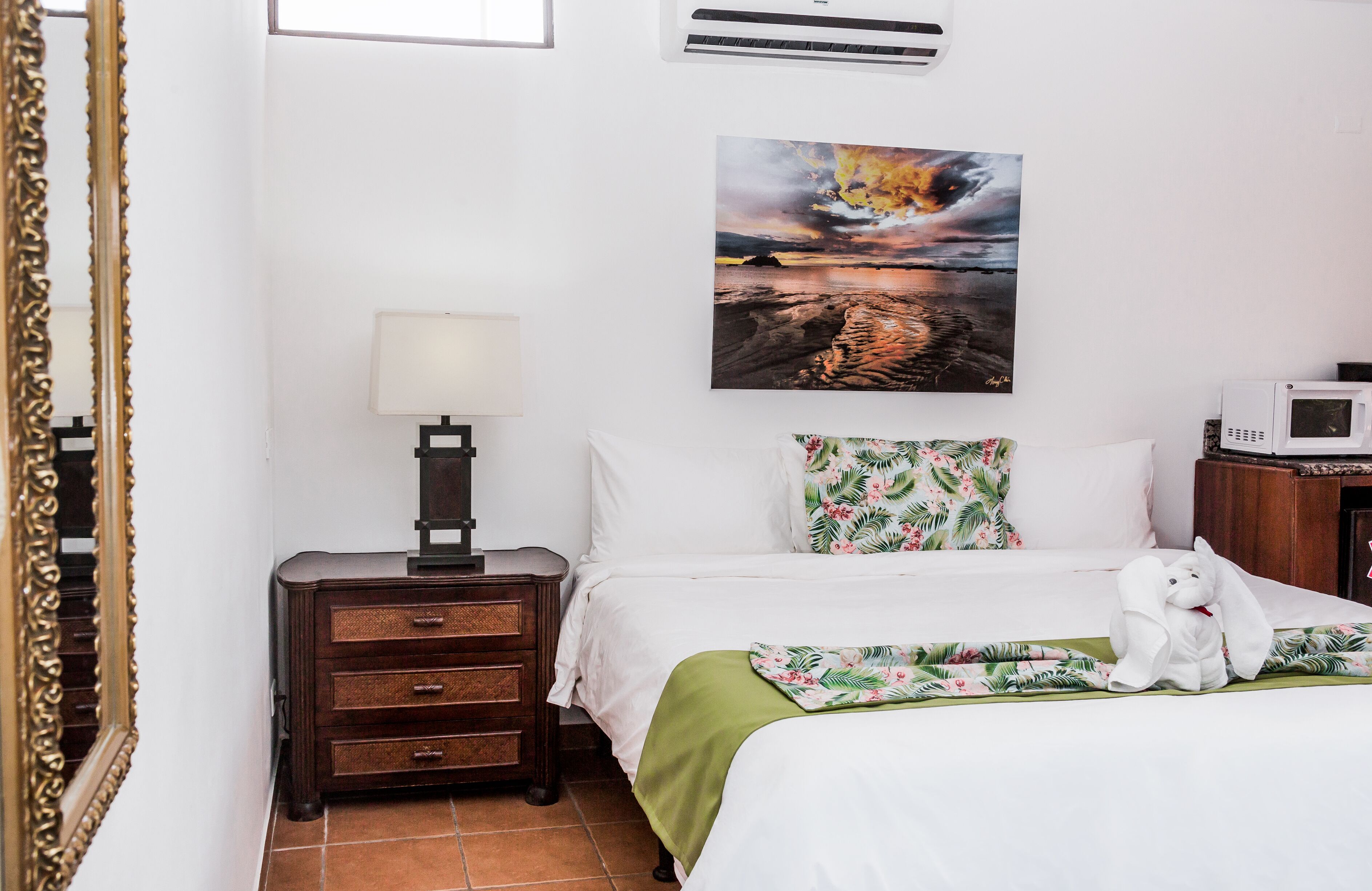 A neatly arranged hotel room with a bed, a nightstand with a lamp, a painting of a sunset, and a microwave on a wooden cabinet.