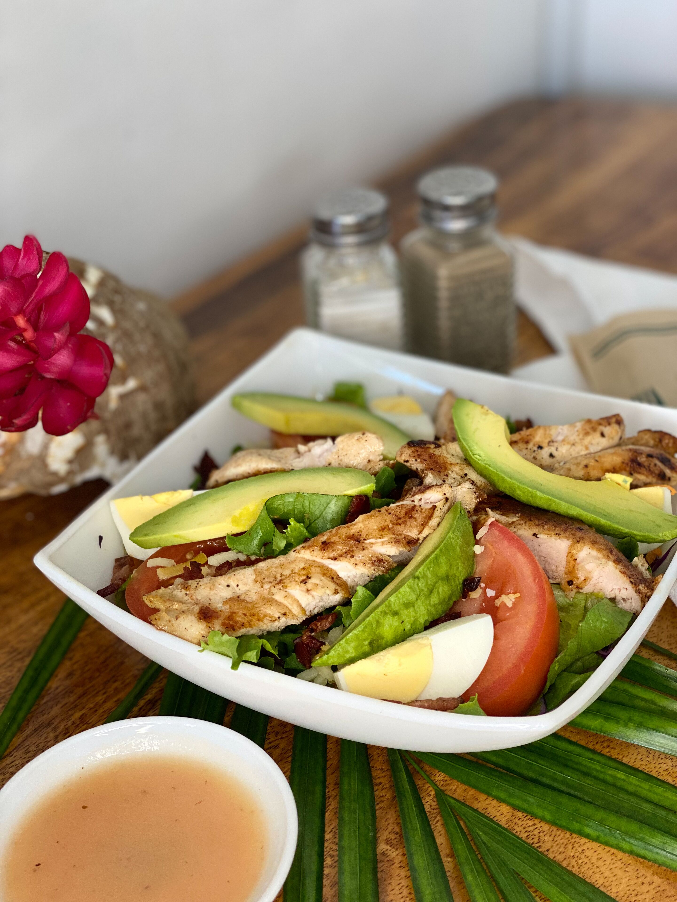 A white bowl of salad with avocado, tomato, and chicken on a wooden table.