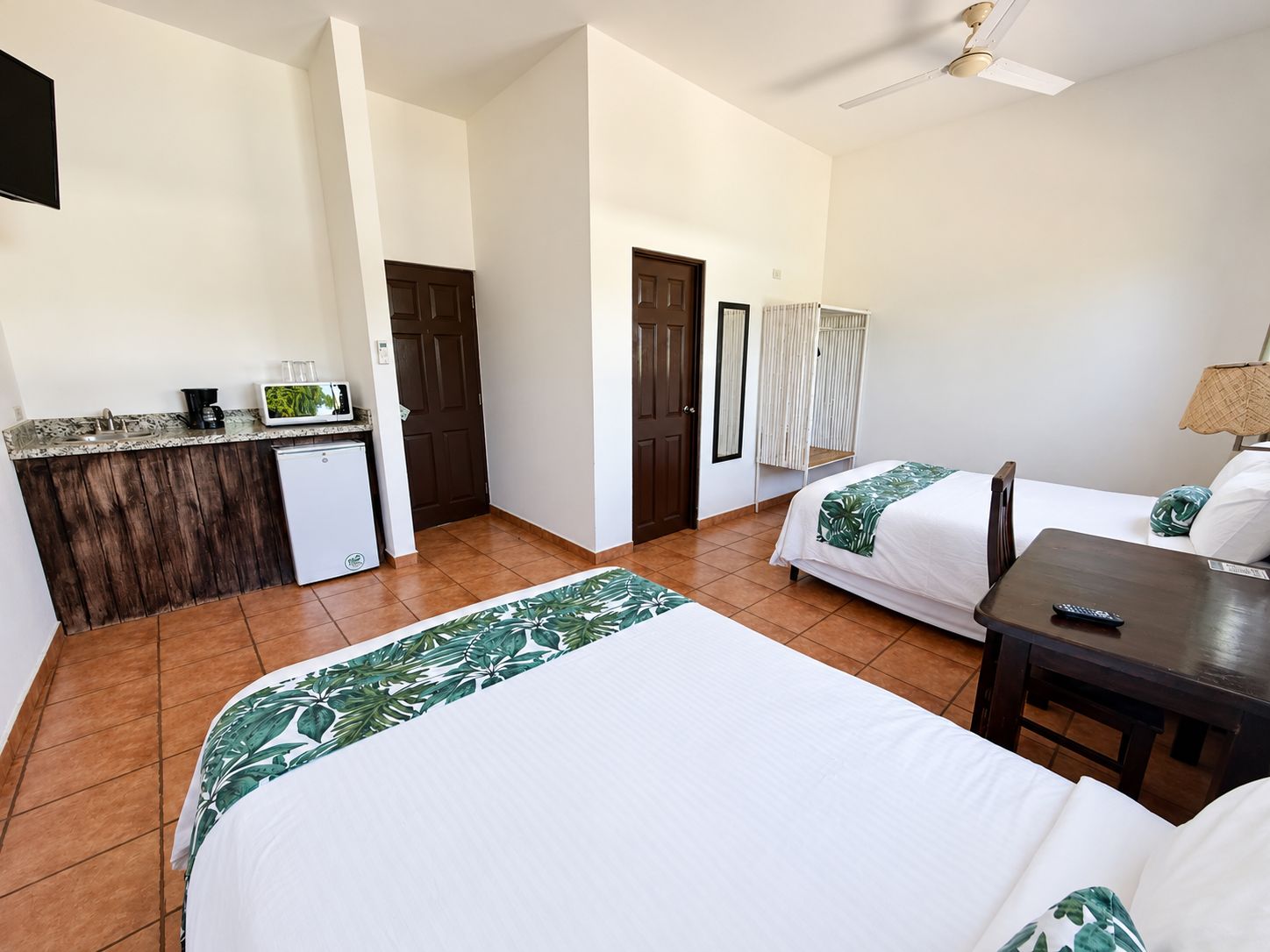 A hotel room with two beds, a small kitchenette, a ceiling fan, and a window.