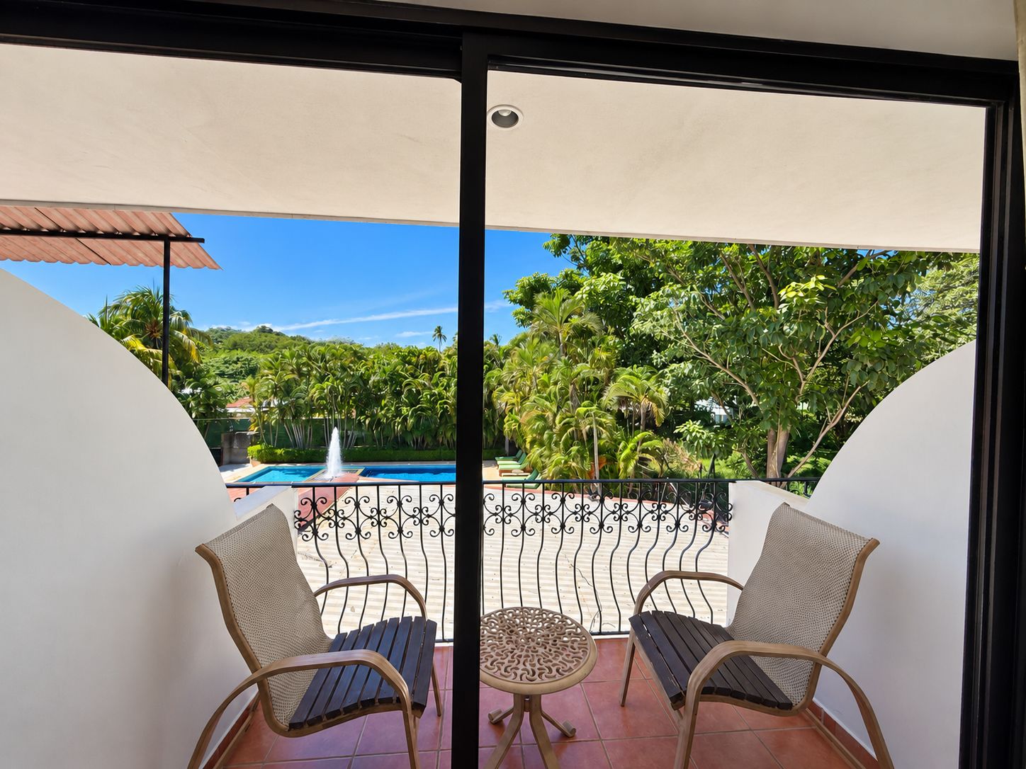 A balcony with two wicker chairs and a small round table, overlooking a swimming pool and lush greenery.