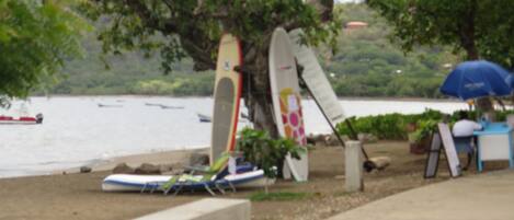A beach with surfboards leaning against a tree and a person sitting under an umbrella.
