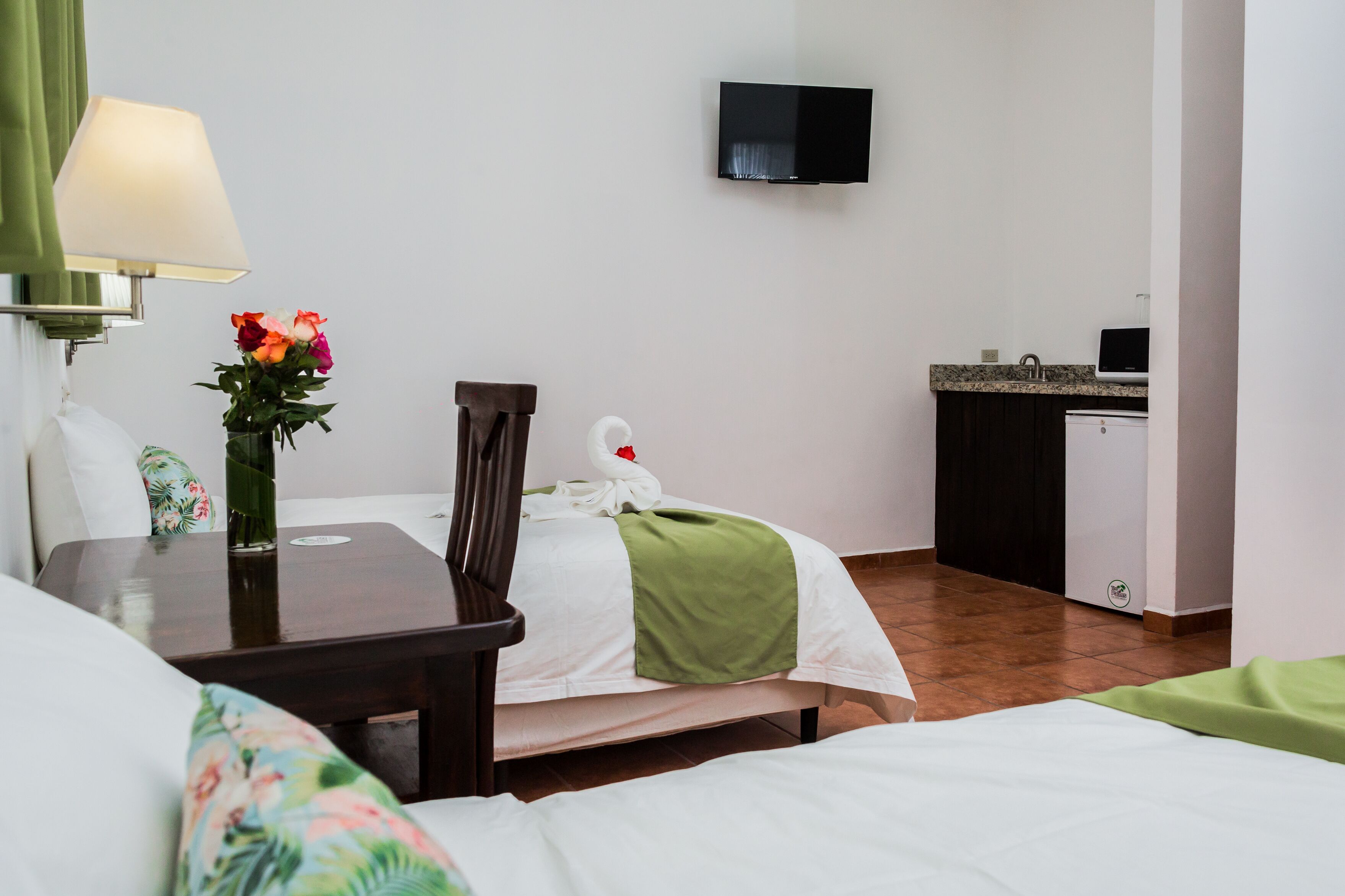 Una habitación de hotel con una cama, un escritorio con lámpara, un florero con flores, un televisor en la pared y una kitchenette.