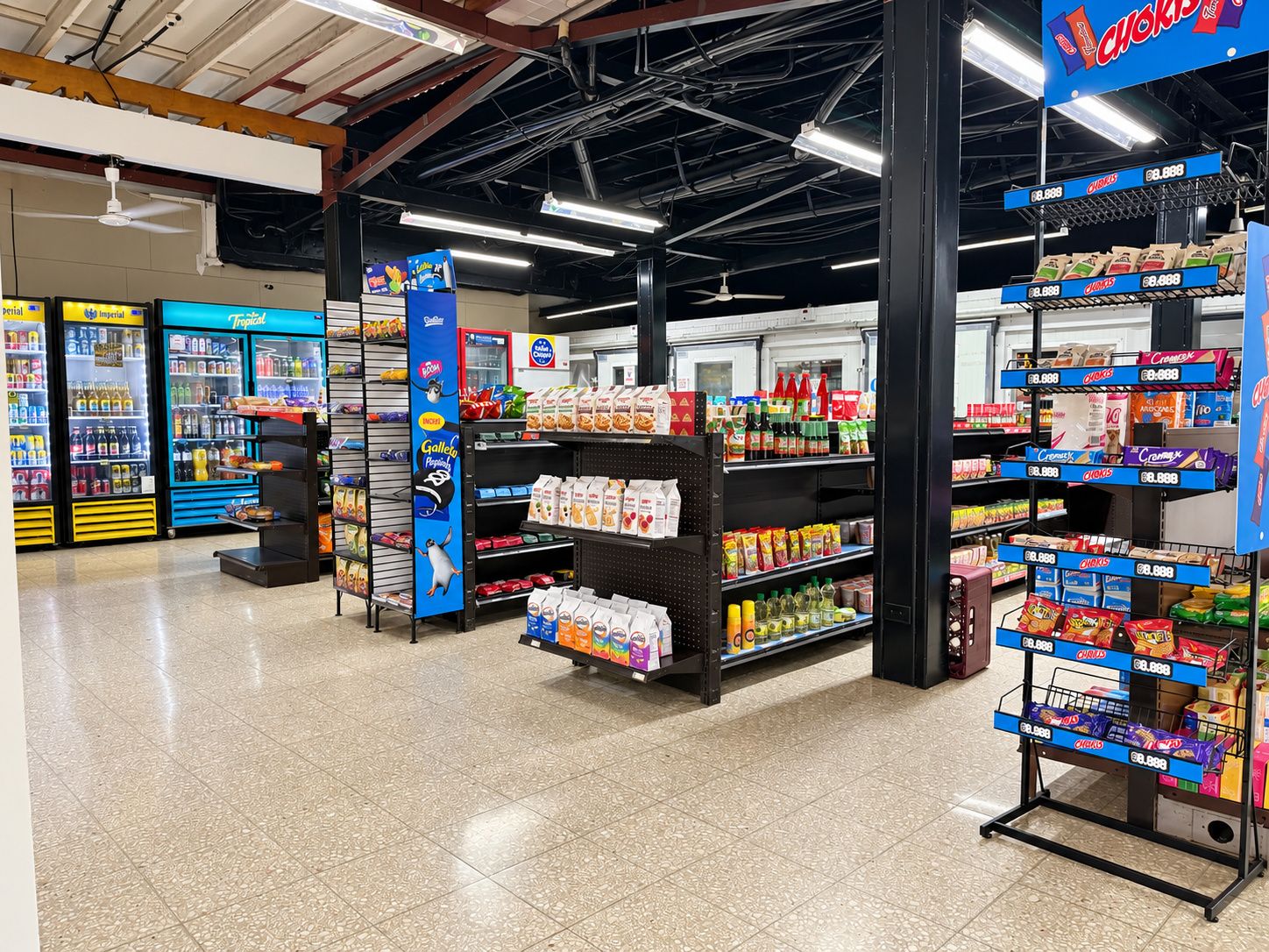 Interior de una tienda de comestibles con secciones refrigeradas, estantes llenos de bocadillos y un techo despejado.