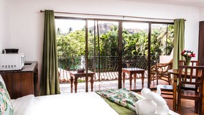 A hotel room with a bed, a desk with a microwave, a balcony with a table and chairs, and a view of greenery and mountains.