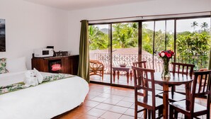 A bedroom with a bed, a nightstand, a microwave, and a Coca-Cola fridge. There is a balcony with a table and chairs, and a view of greenery.