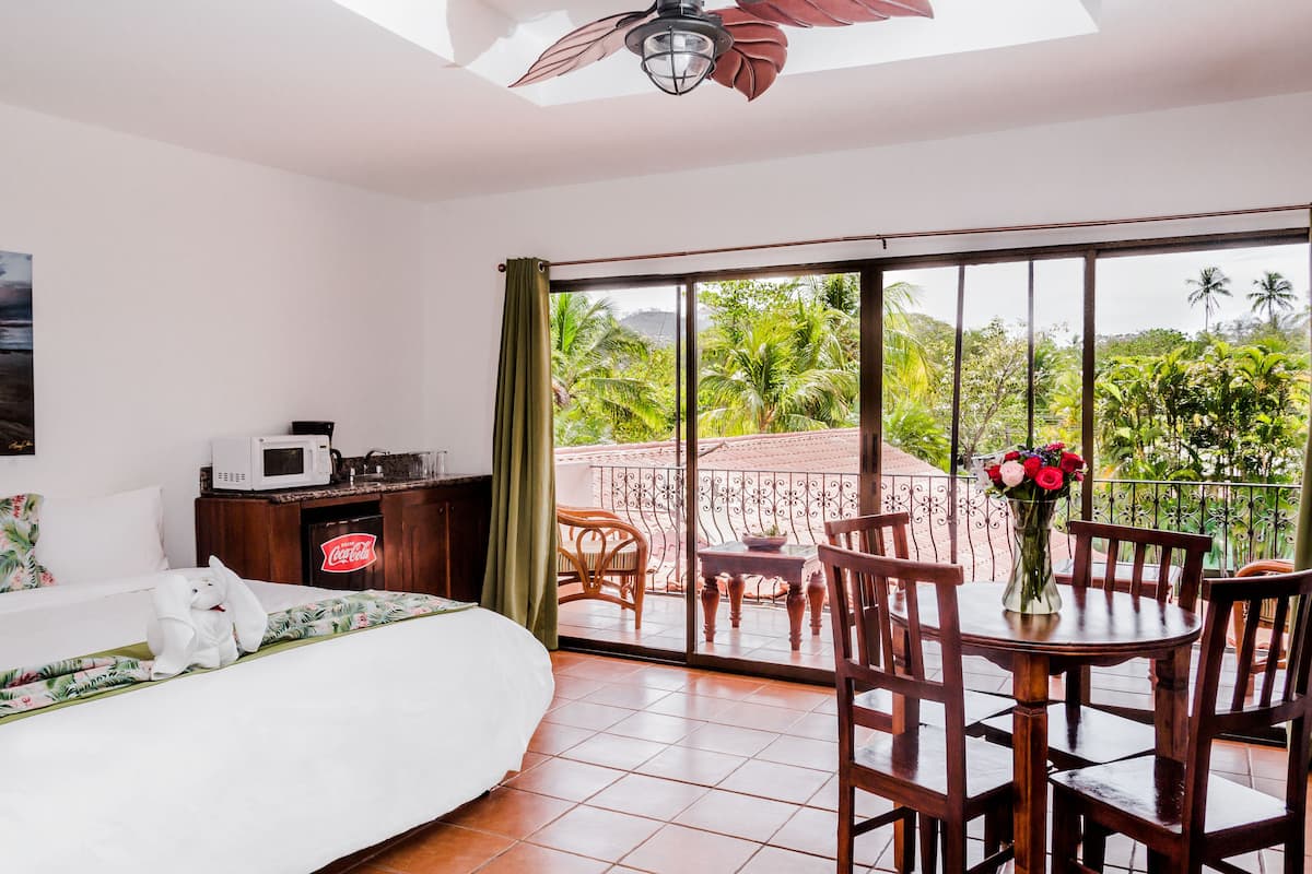 A bedroom with a bed, a nightstand, a microwave, and a Coca-Cola fridge. There is a balcony with a table and chairs, and a view of greenery.