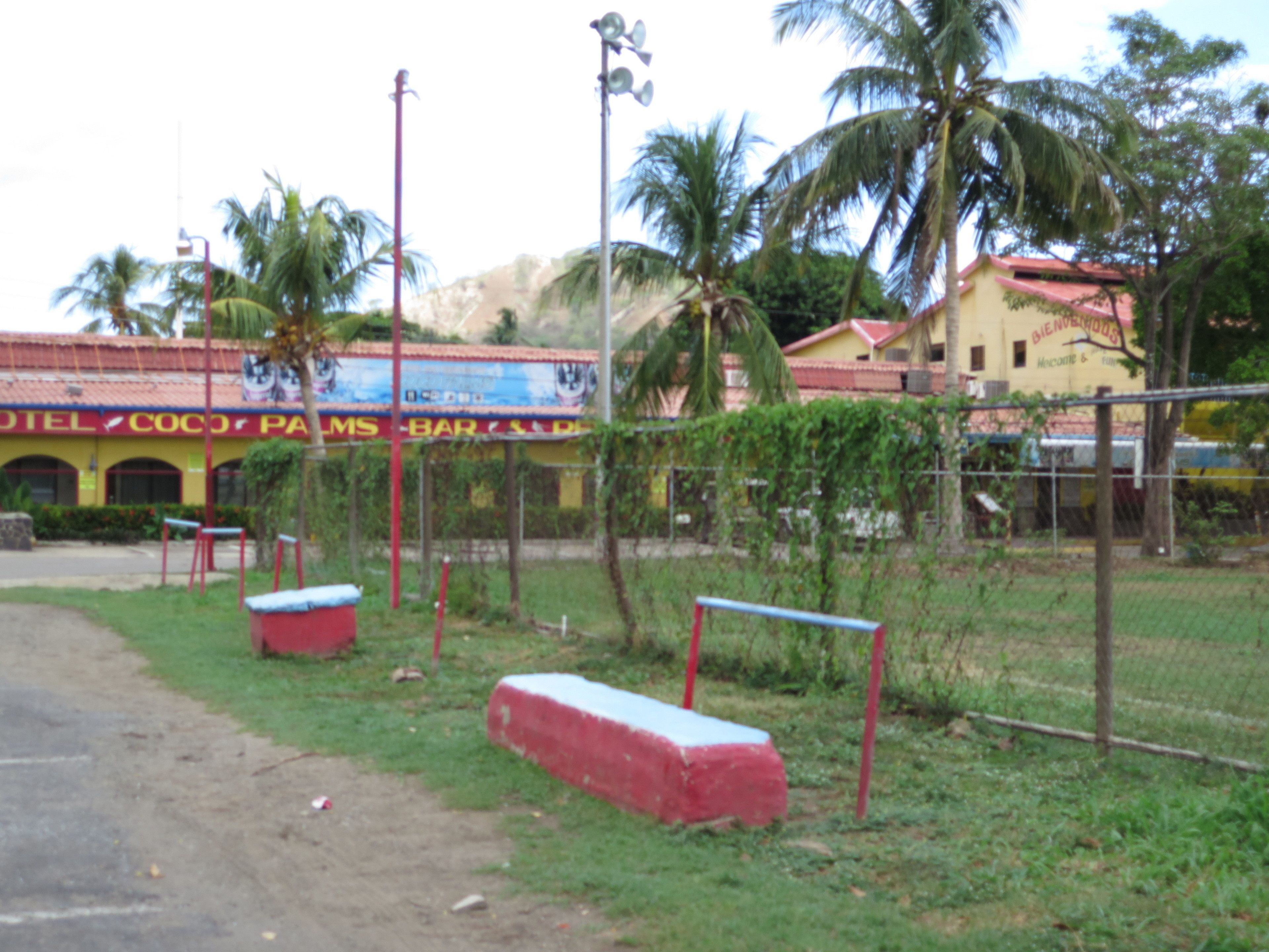 A hotel with a red and yellow facade, palm trees, and a fenced outdoor area with benches.